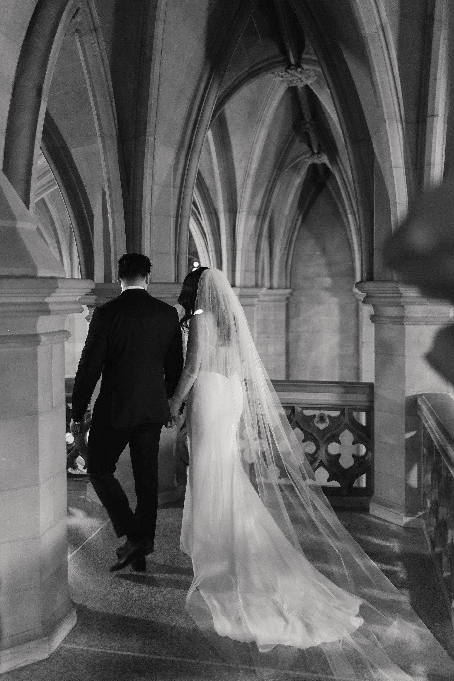 Black and white photo of newlyweds walking through a cathedral hallway with flowing veil trailing behind.