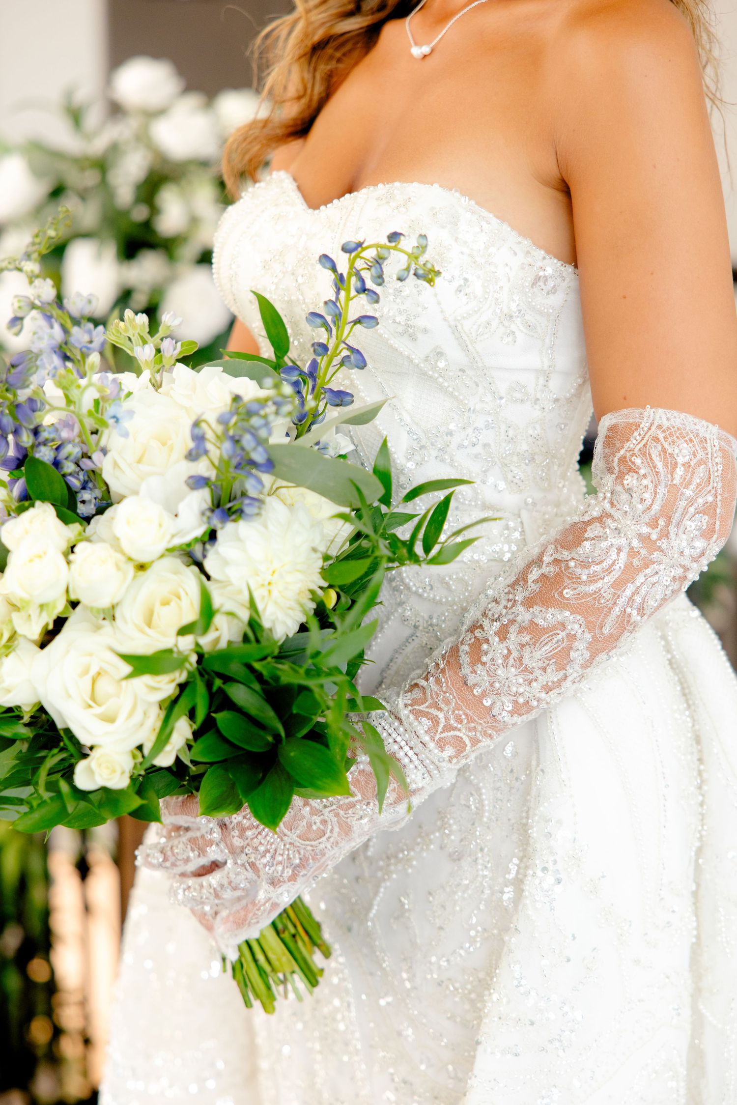 Elegant bridal bouquet with white roses and blue delphinium flowers against a detailed lace wedding dress.