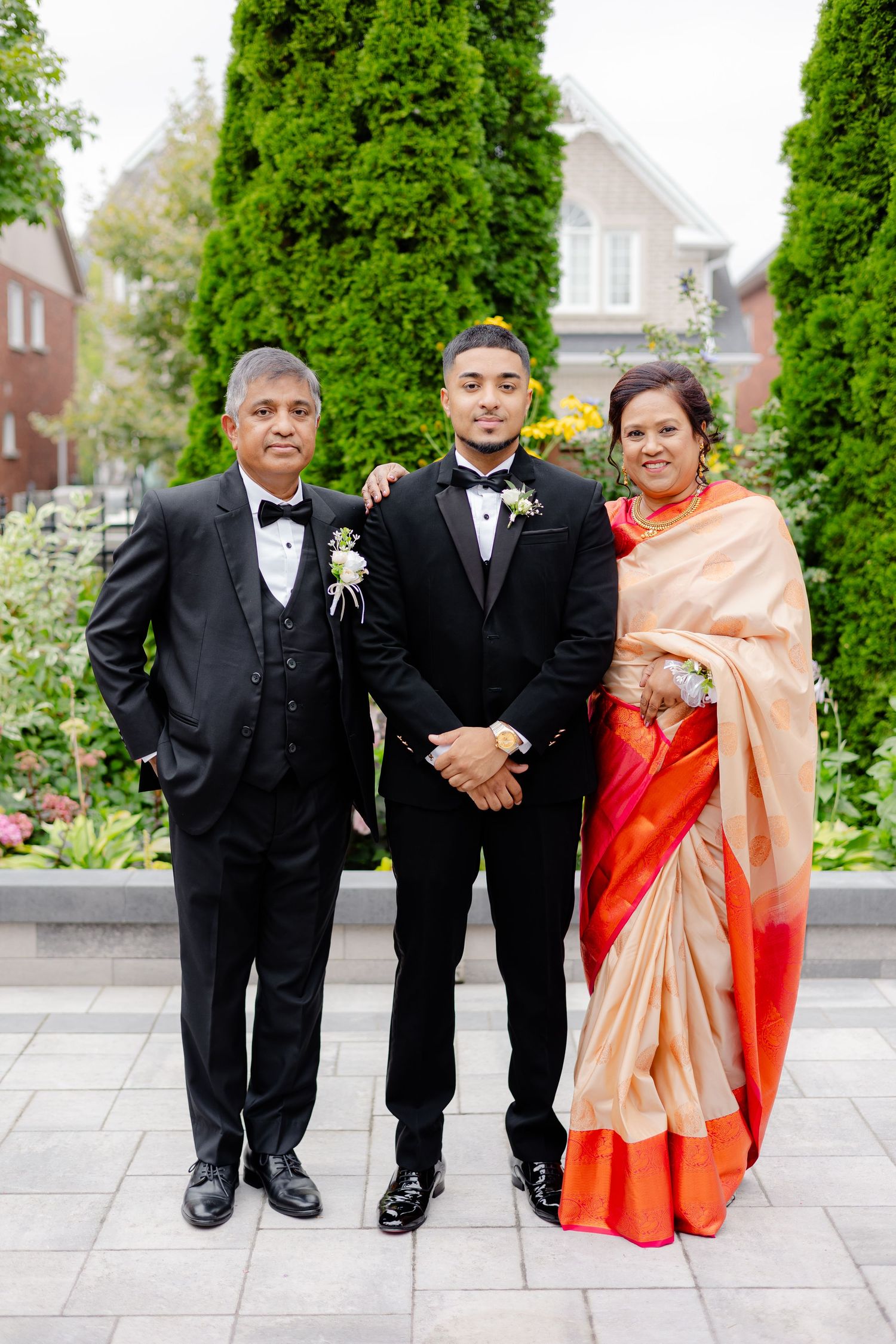 Formal wedding portrait in front of house with decorative evergreen trees.