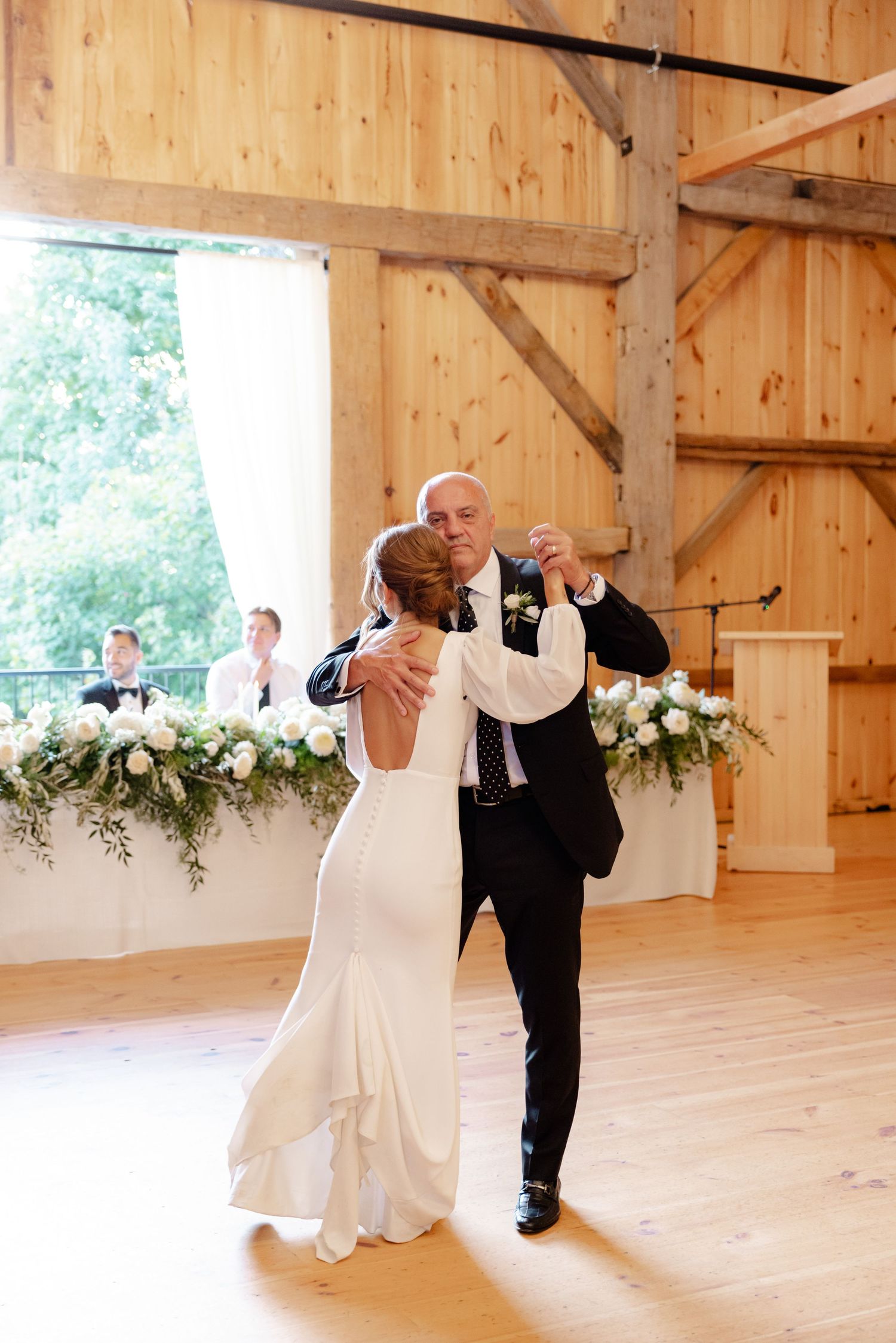 A couple dances together in a sunlit barn with elegant floral decorations.