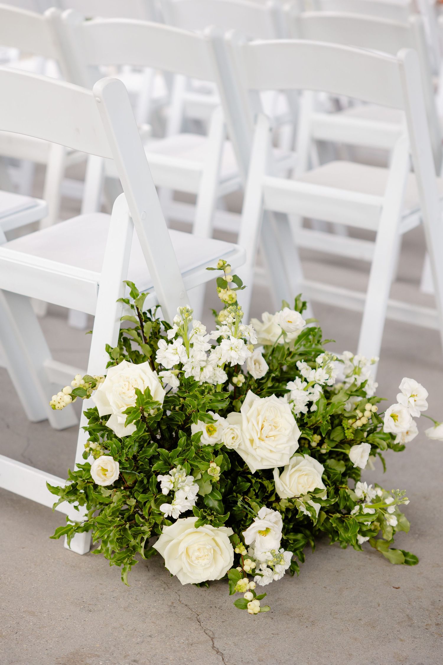 White roses and greenery arranged as wedding ceremony aisle decorations beside folding chairs.