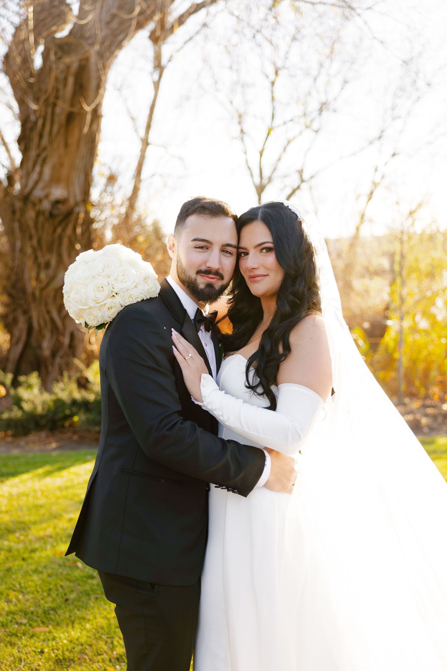 Elegant wedding portrait with couple embracing in front of rustic tree with white bouquet.