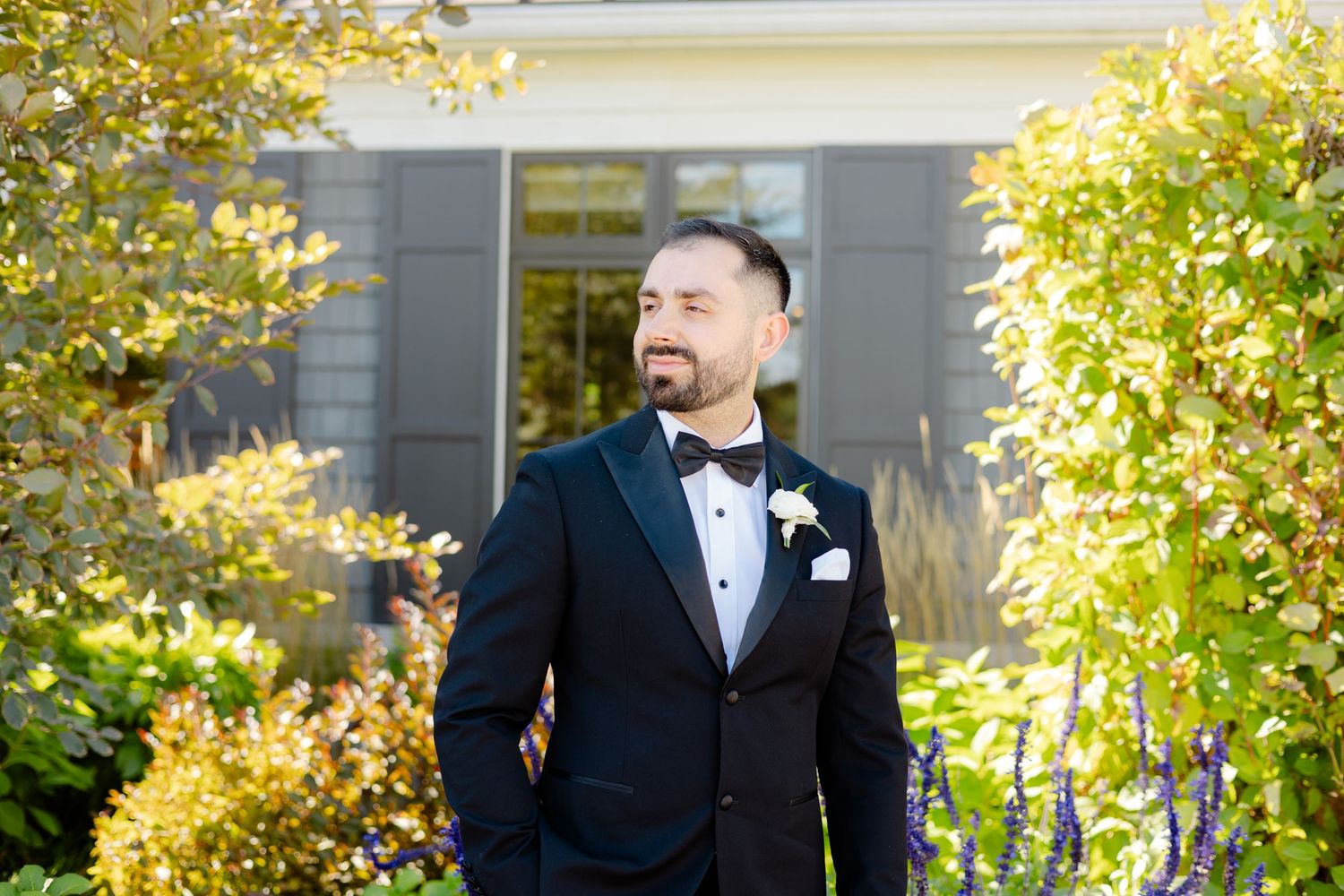 Groom in classic tuxedo smiling in front of house with autumn foliage.