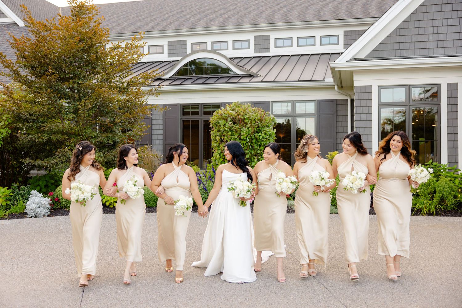 Wedding party members in cream-colored dresses walk together outside an elegant gray building.