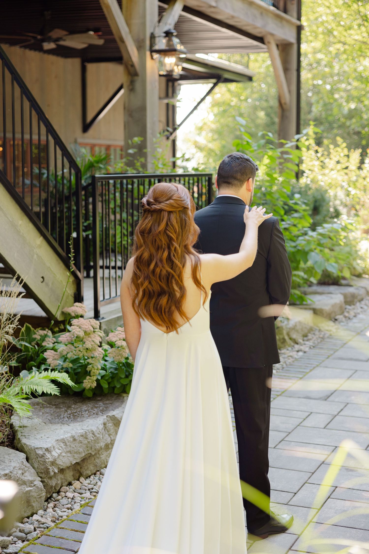 Bride approaches groom from behind on stone path under rustic wooden overhang.