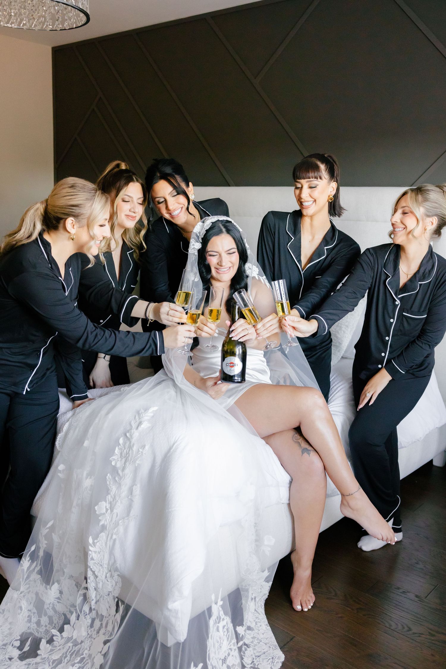 Wedding party laughs while popping champagne bottle during pre-ceremony celebration.