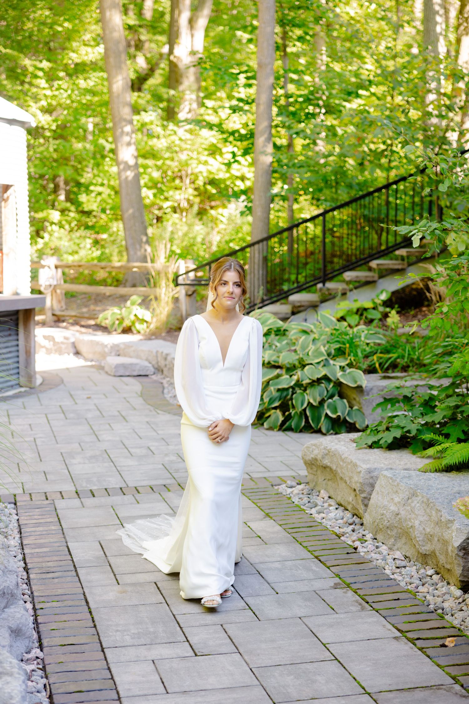 Elegant bride in a fitted white gown poses on a paved pathway surrounded by natural landscaping.