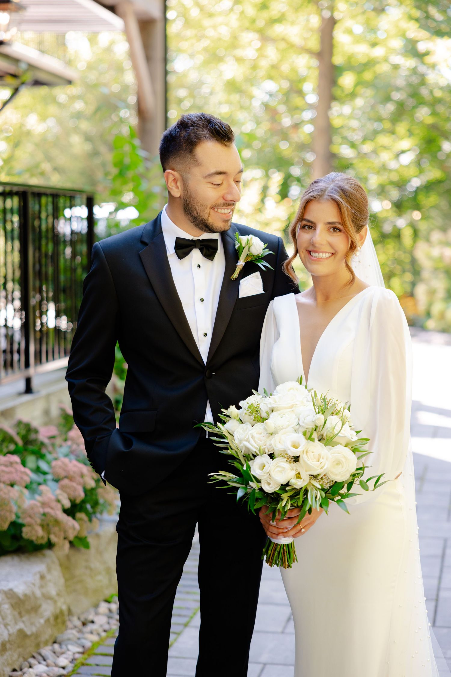 A happy couple in wedding attire poses together outdoors surrounded by lush greenery.