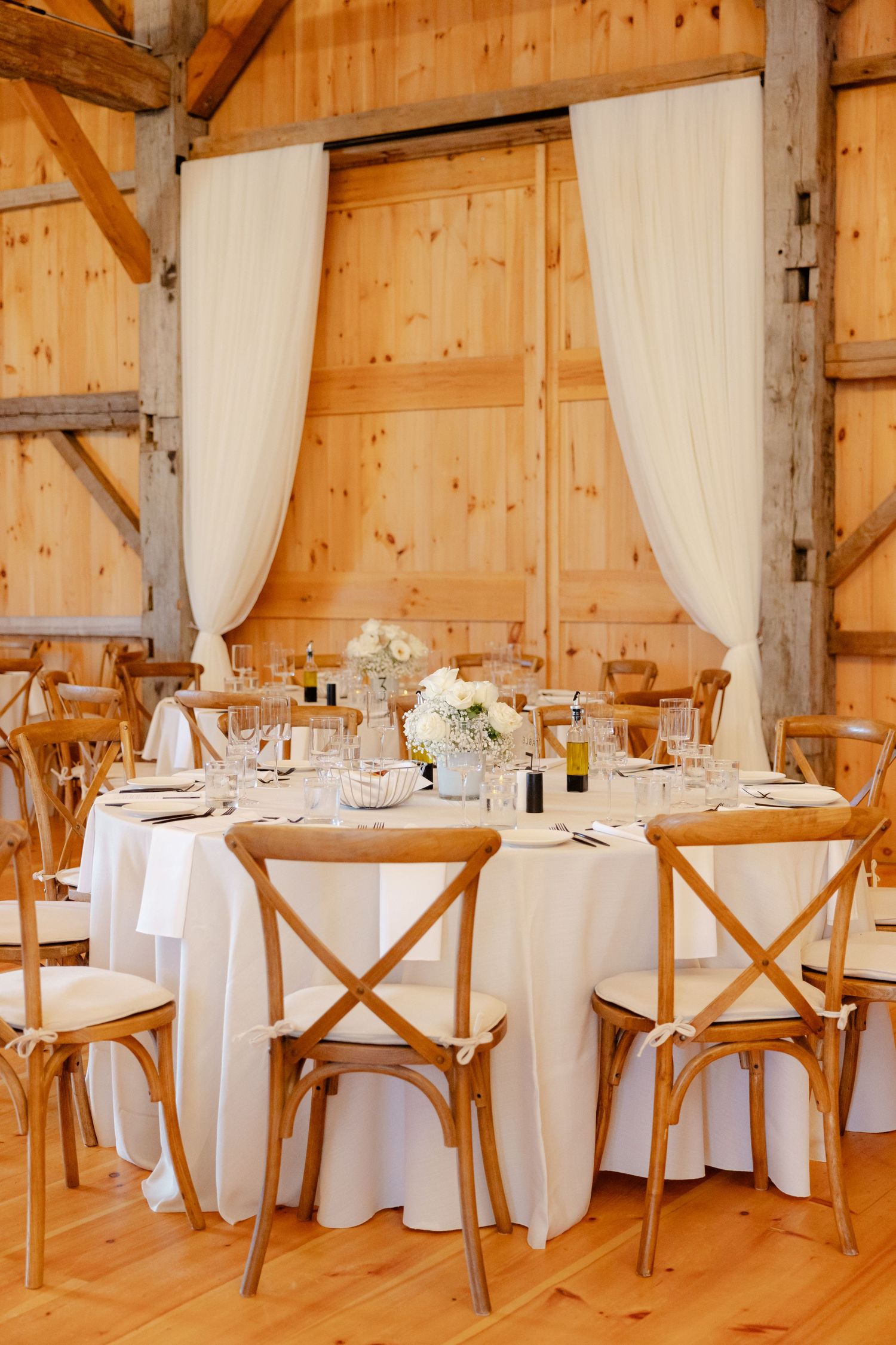 Elegant barn wedding reception setup with round tables, white linens, and rustic crossback chairs in a wooden venue.