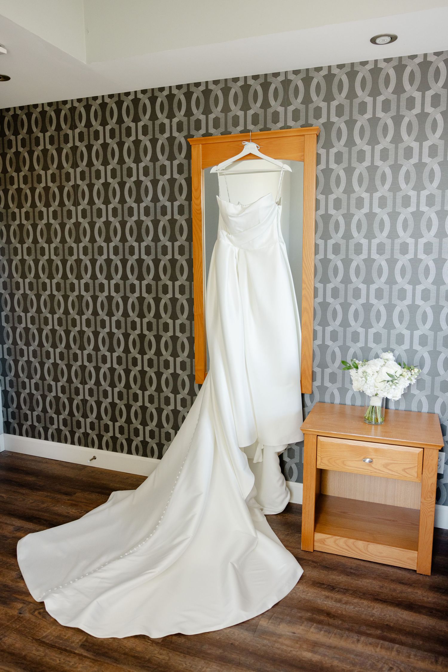 Elegant white wedding dress with long train hanging against gray patterned wallpaper next to wooden nightstand.