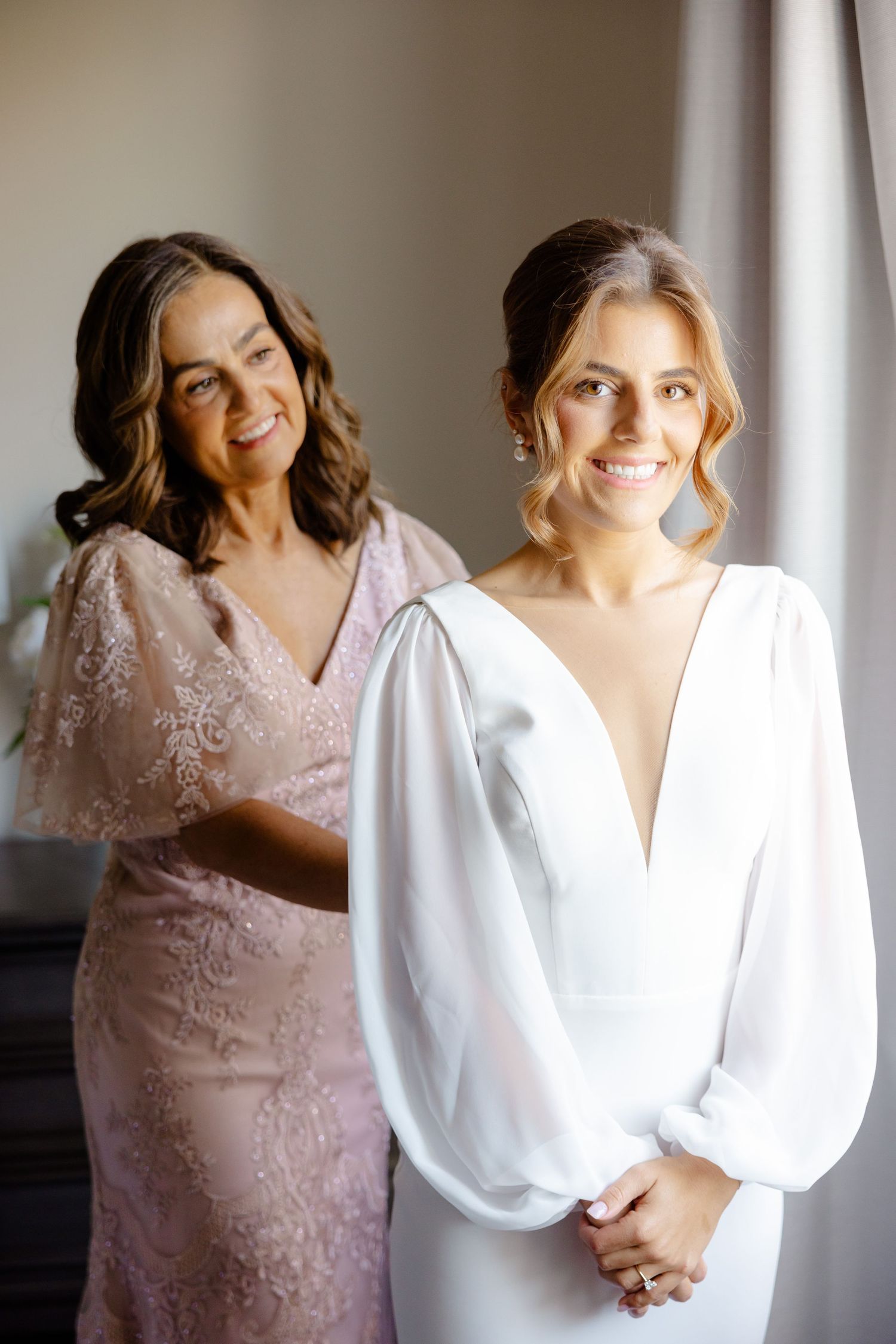Two people pose together wearing formal dresses in white and blush pink near a window.