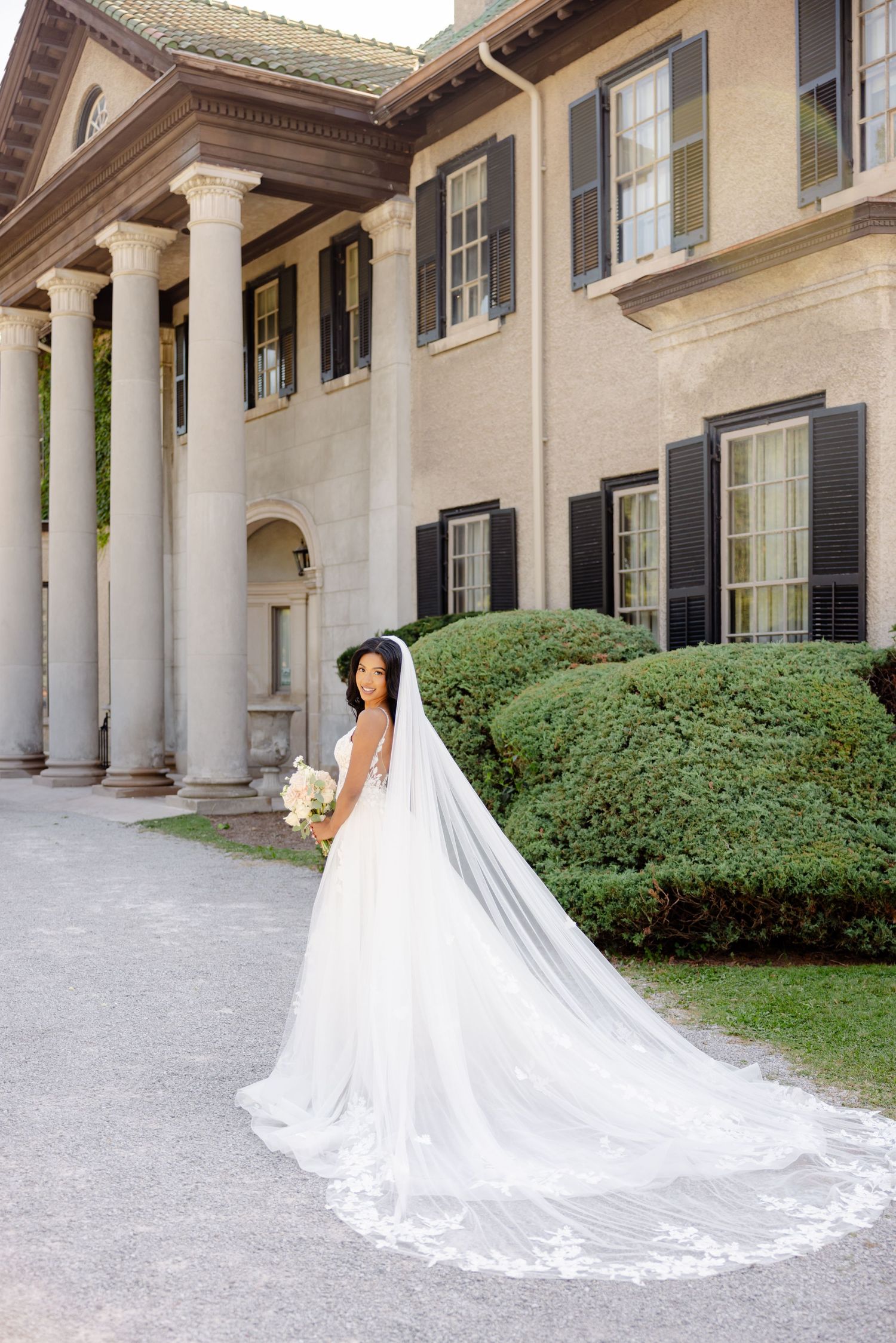 Bride with flowing cathedral veil stands against backdrop of historic mansion with columns.