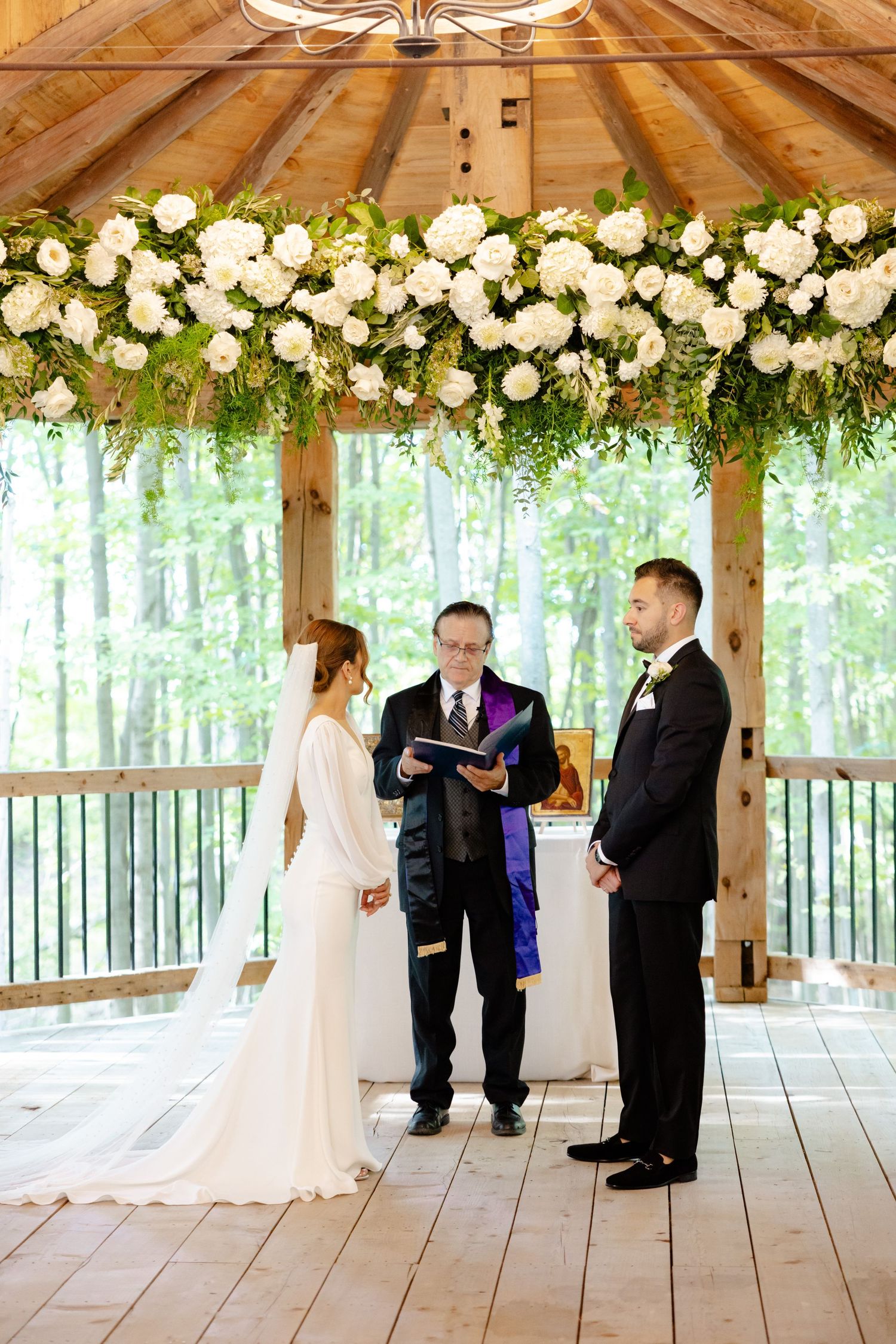 A wedding ceremony under a floral arch with white roses in a wooden pavilion.