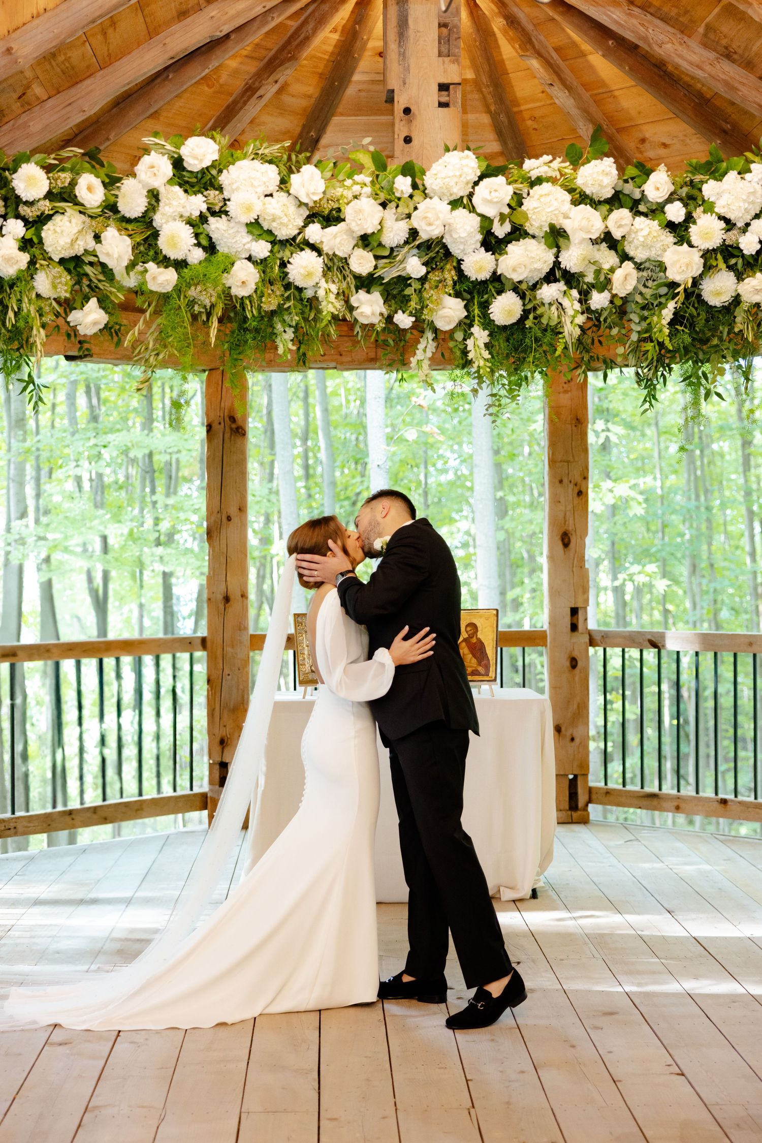 A wedding couple embraces under a wooden gazebo decorated with white flowers and greenery in a forest setting.