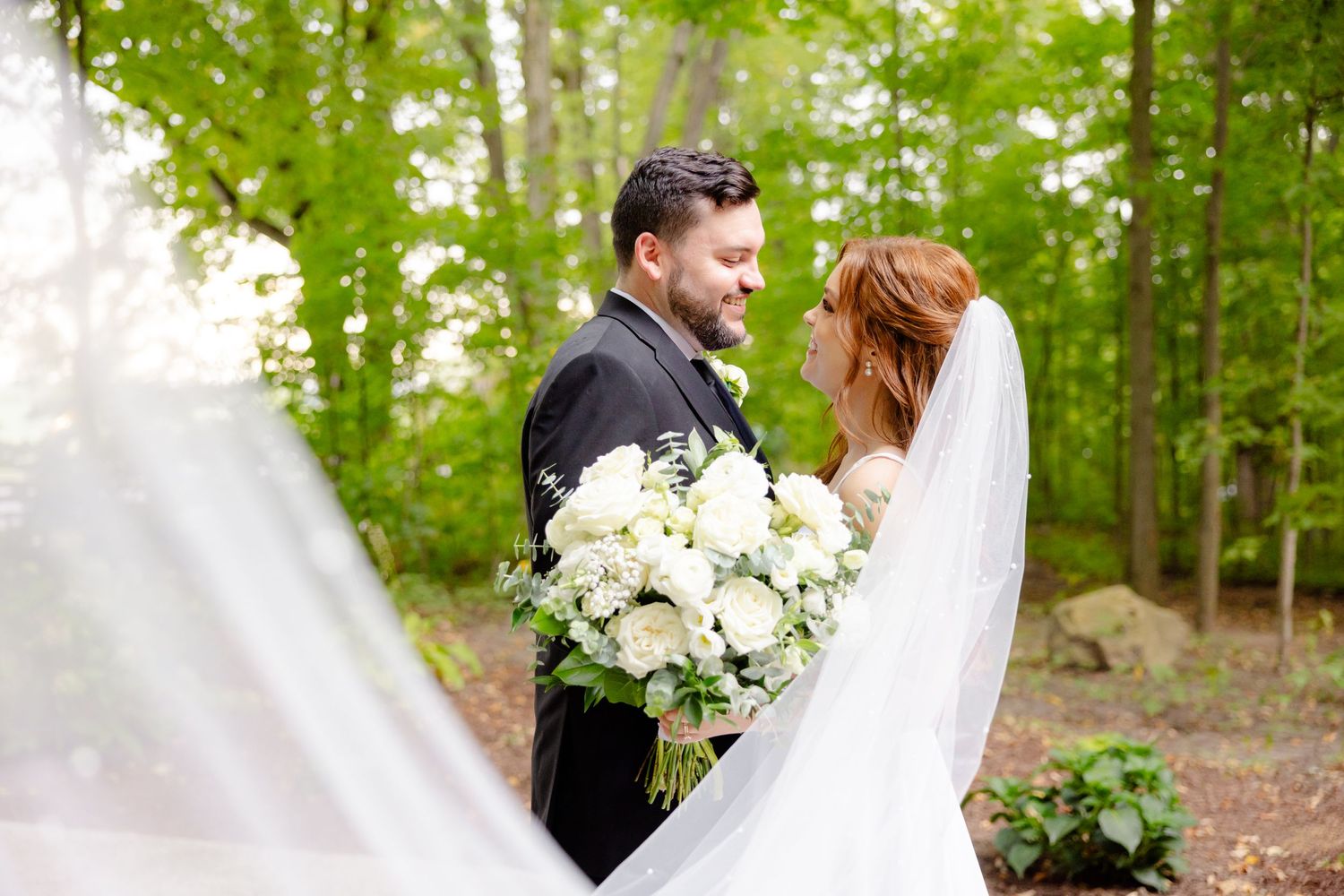 A romantic wedding moment captured in a sunlit wooded setting with white floral bouquet.