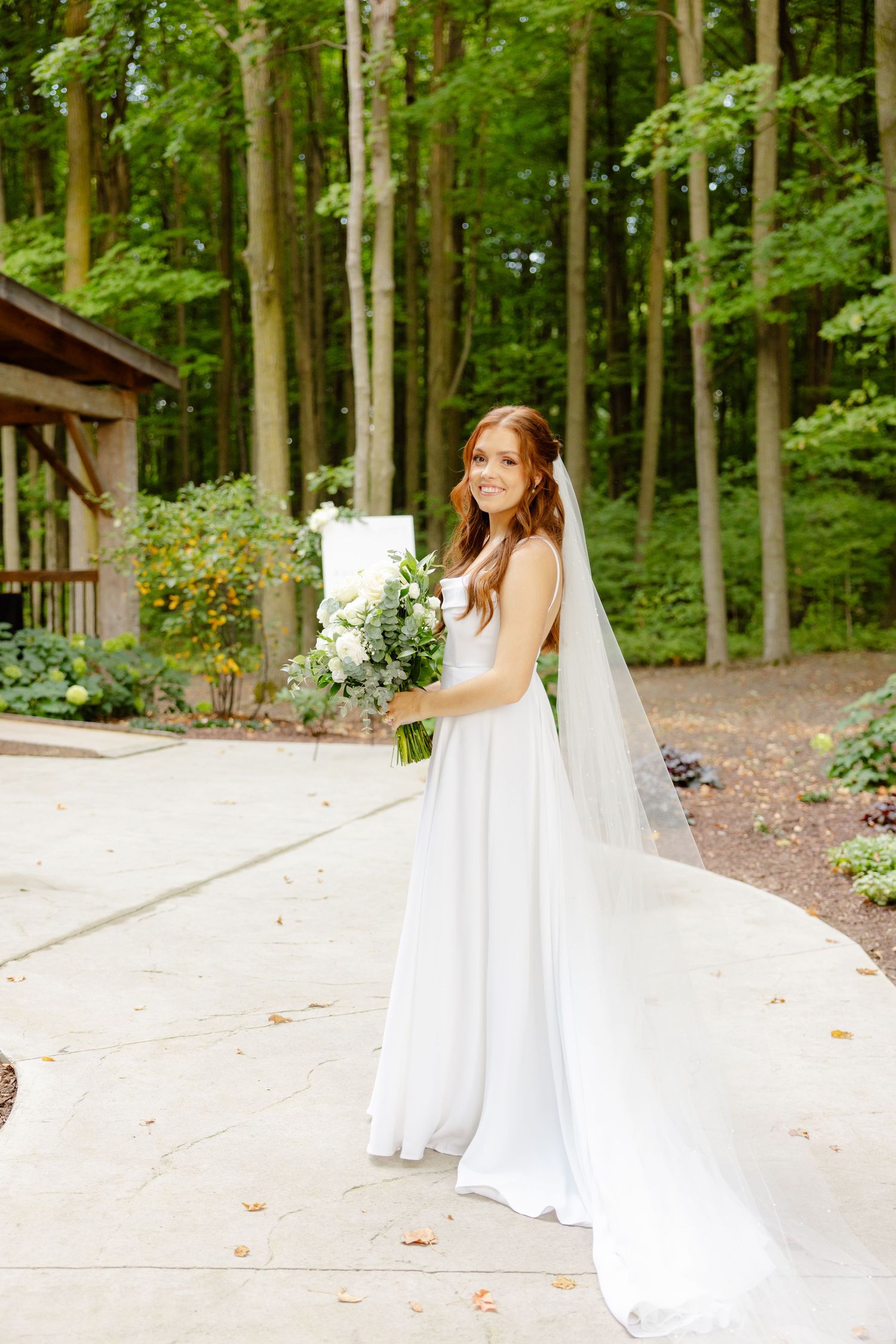Bride in white gown with flowing veil holds bouquet while standing on concrete path near forest pavilion.