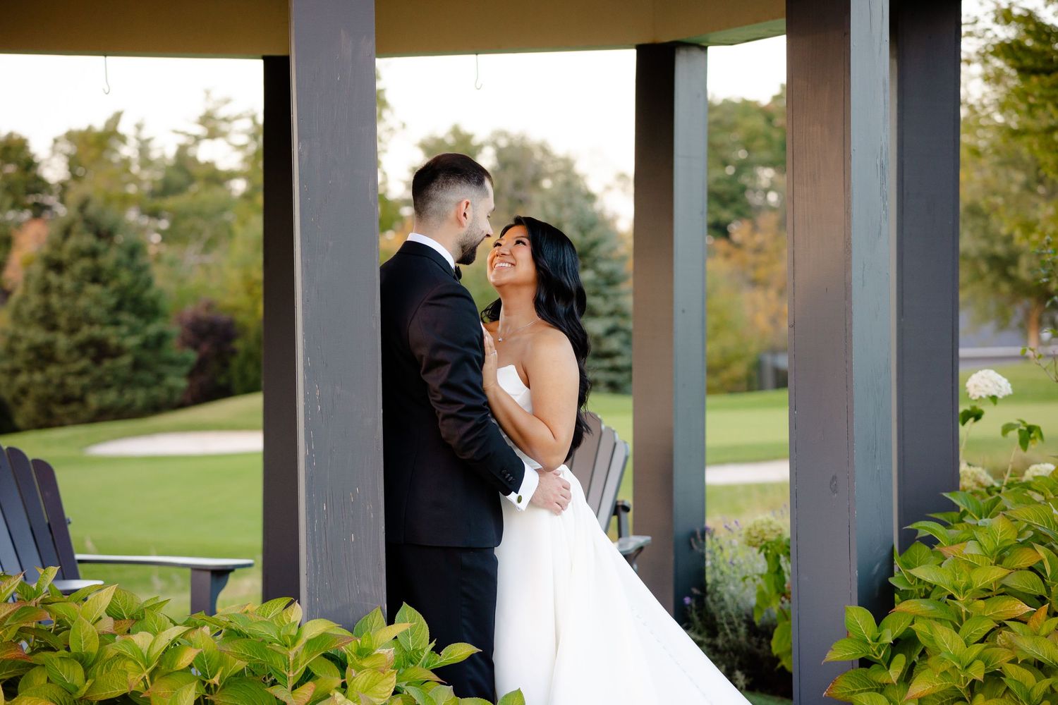 A romantic wedding couple embraces under an outdoor gazebo at sunset with lush greenery in the background.