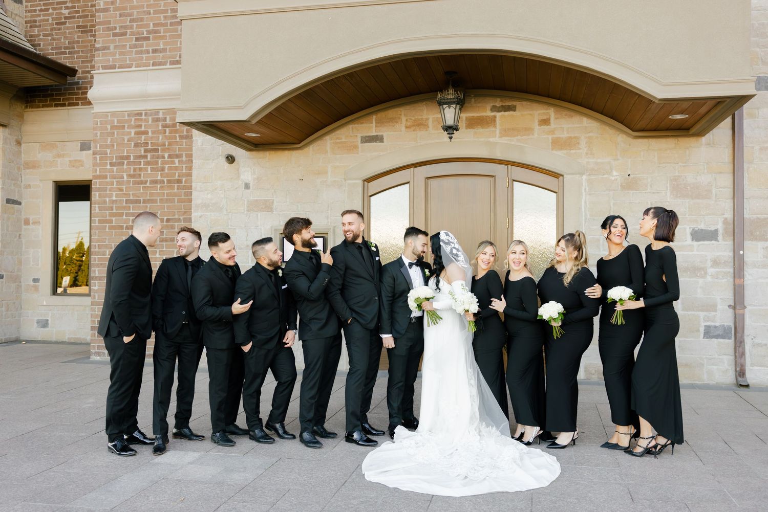 Wedding party shares a candid moment outside stone building with bride in white gown surrounded by attendants in black.