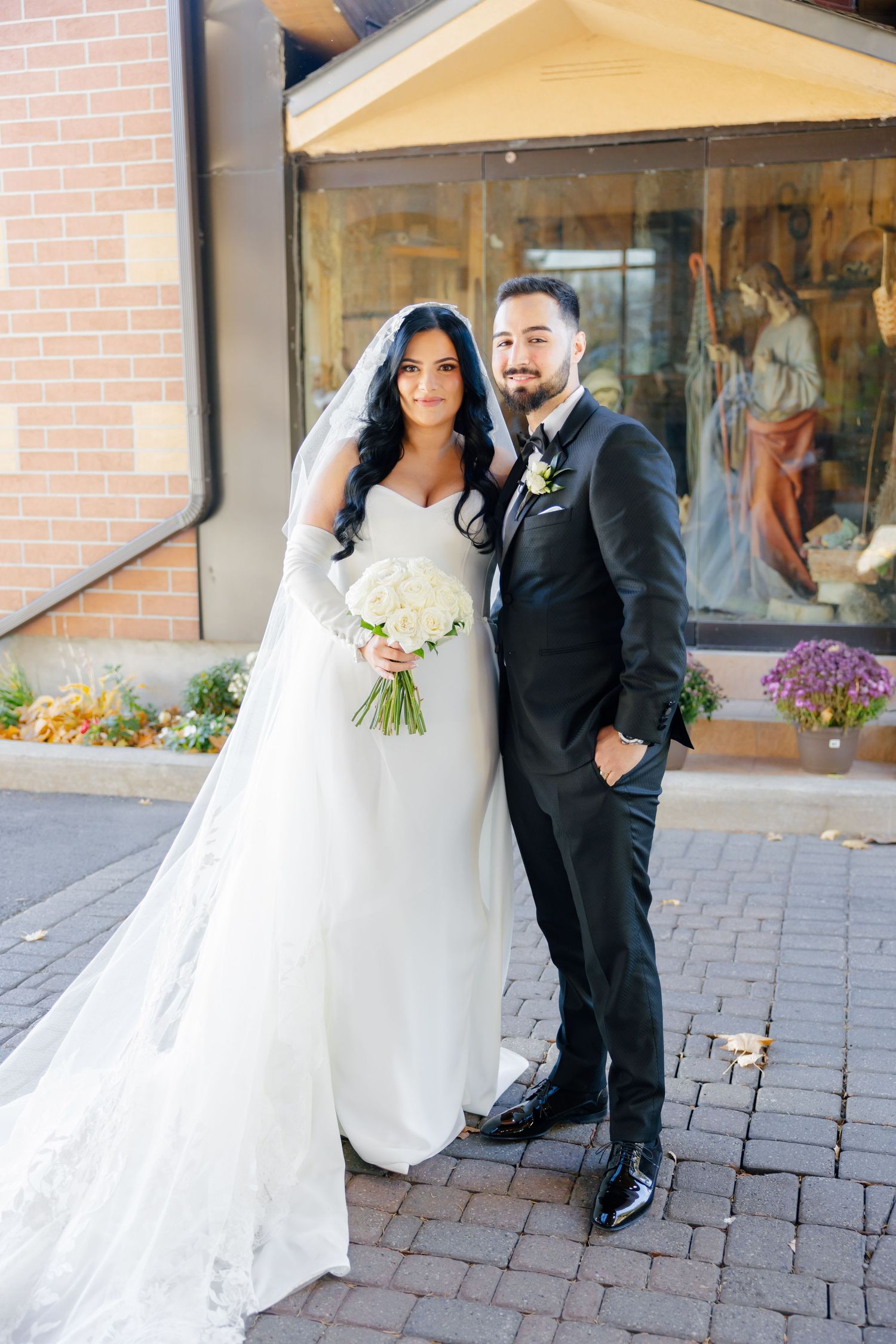 A newly married couple poses outside a brick building with the bride holding a white bouquet.