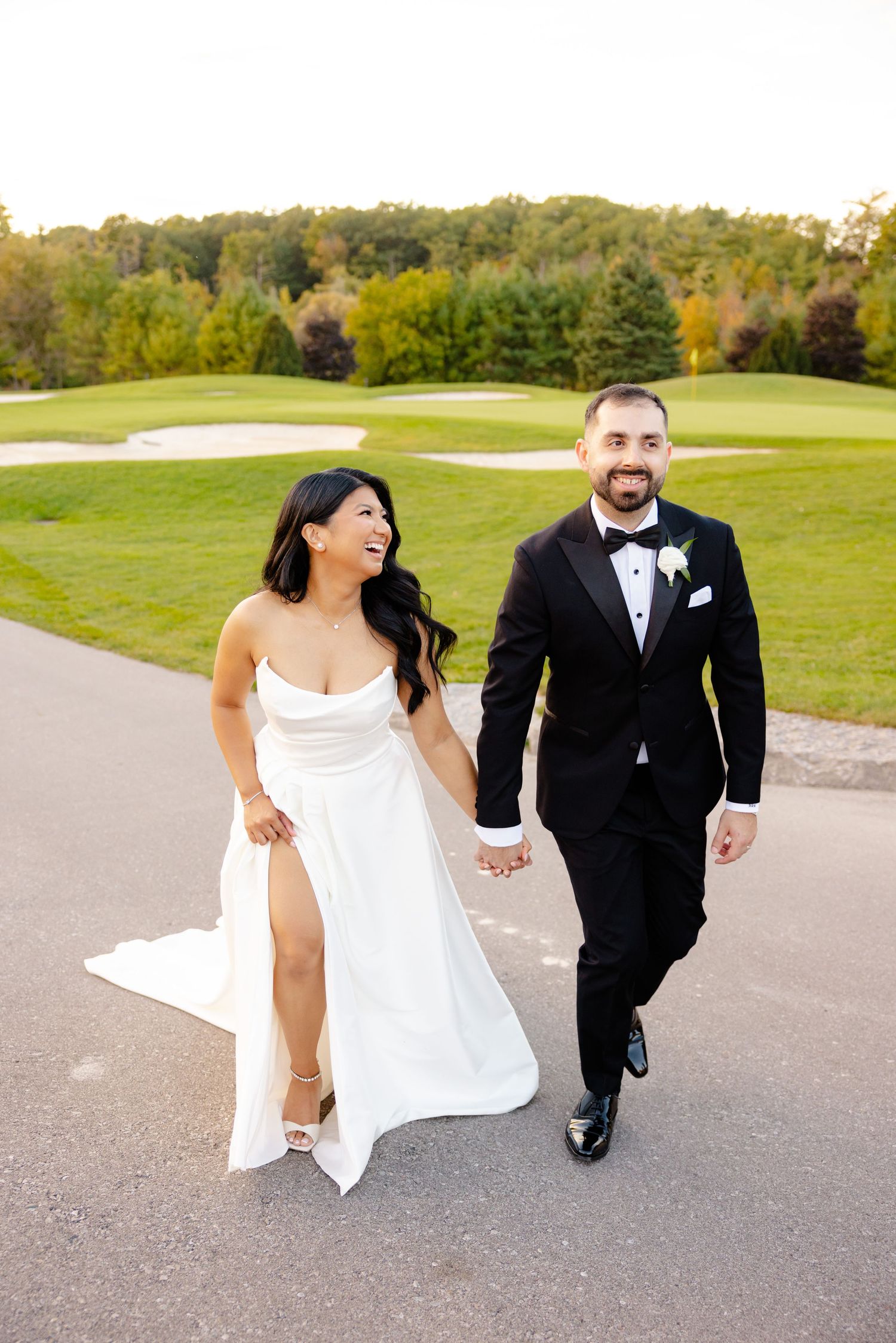 Couple walks hand in hand on golf course path, bride's dress flowing in elegant slit design.