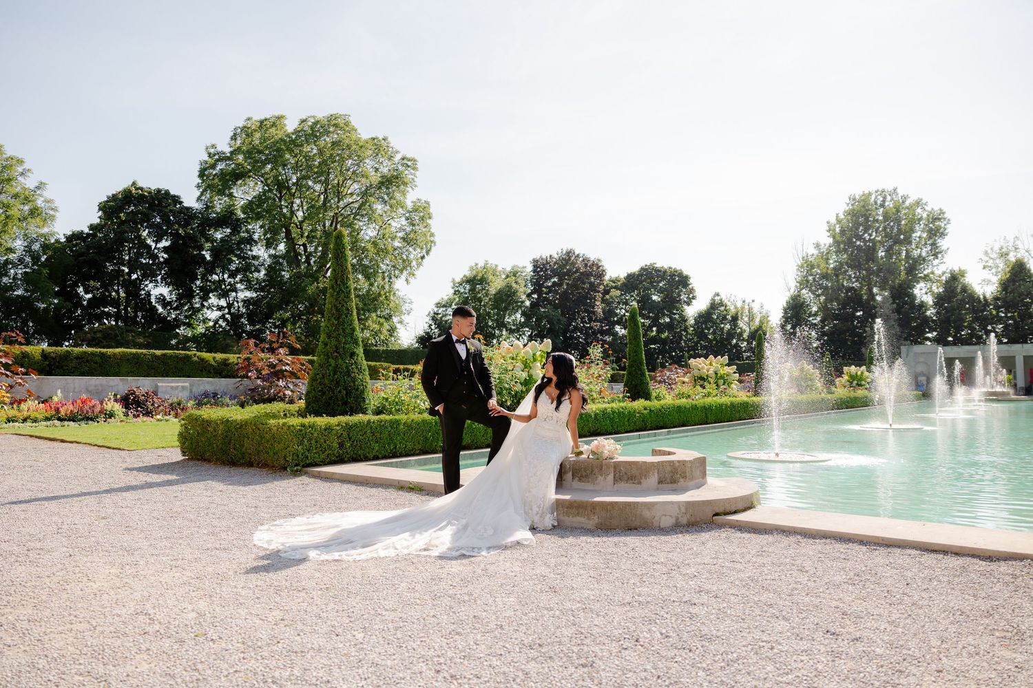 A couple in wedding attire walks near a fountain in a beautifully landscaped garden.