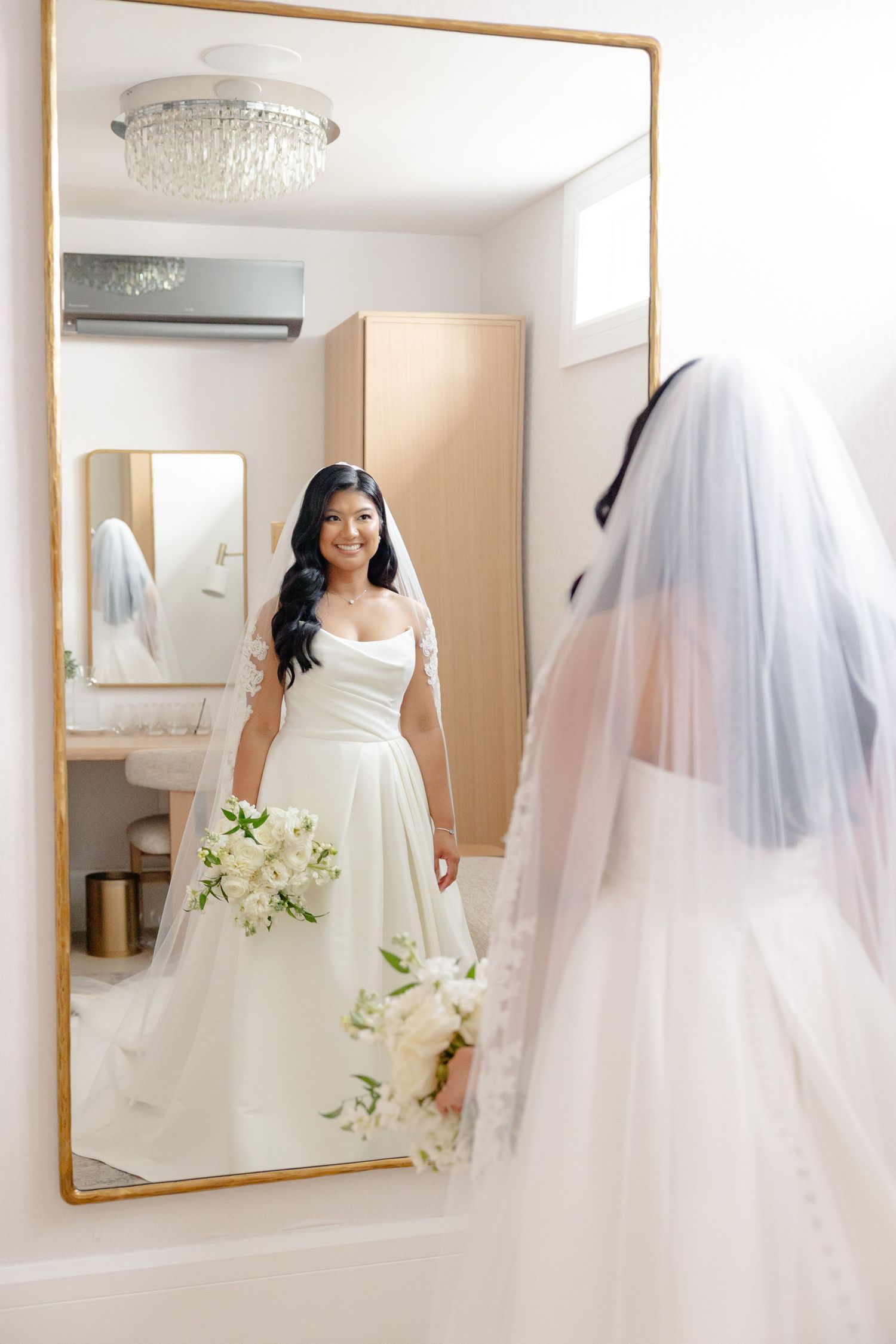Bride in white gown looks at reflection in full-length mirror while holding bouquet.