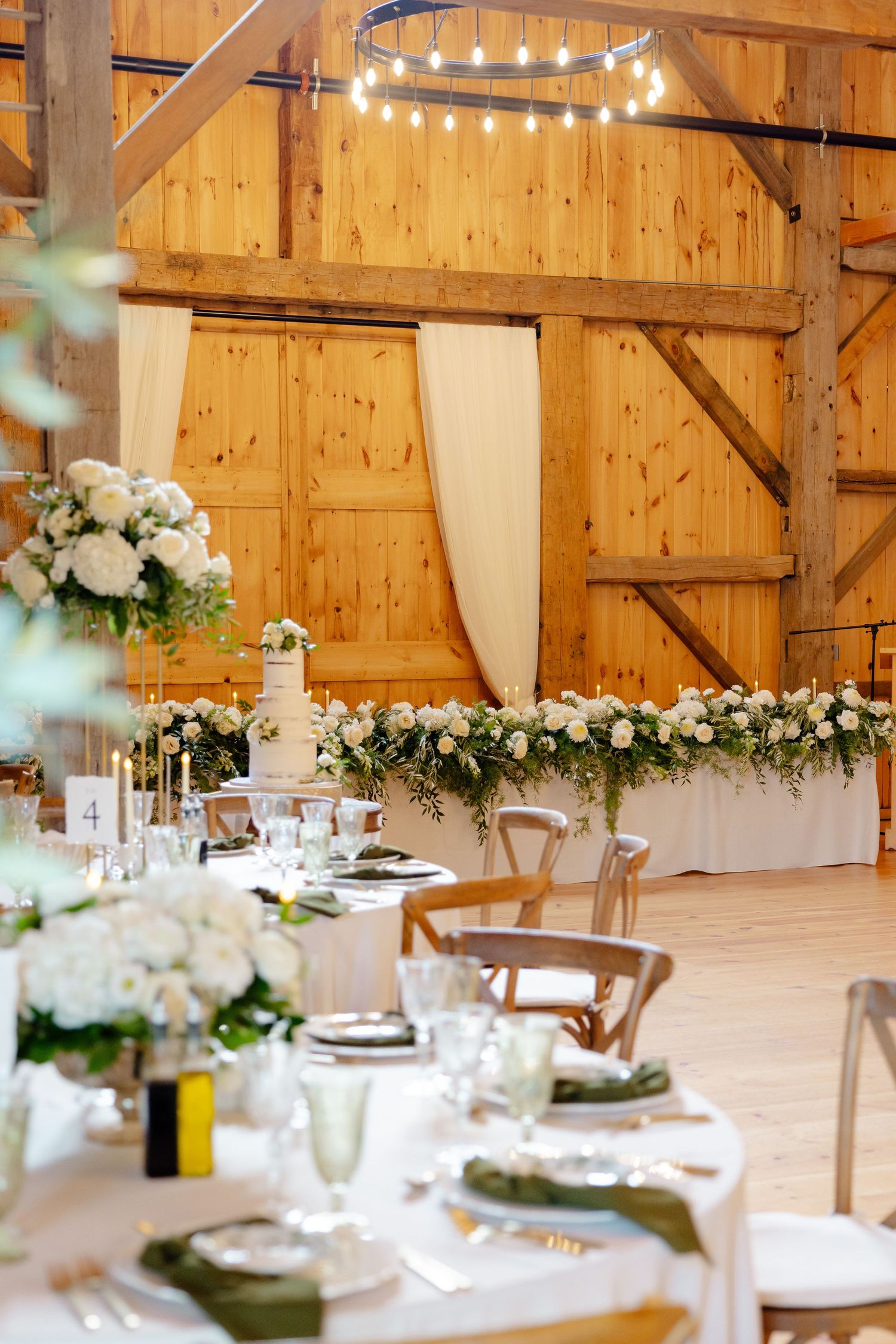 Wedding reception setup in a barn with string lights, white linens, and floral decorations.