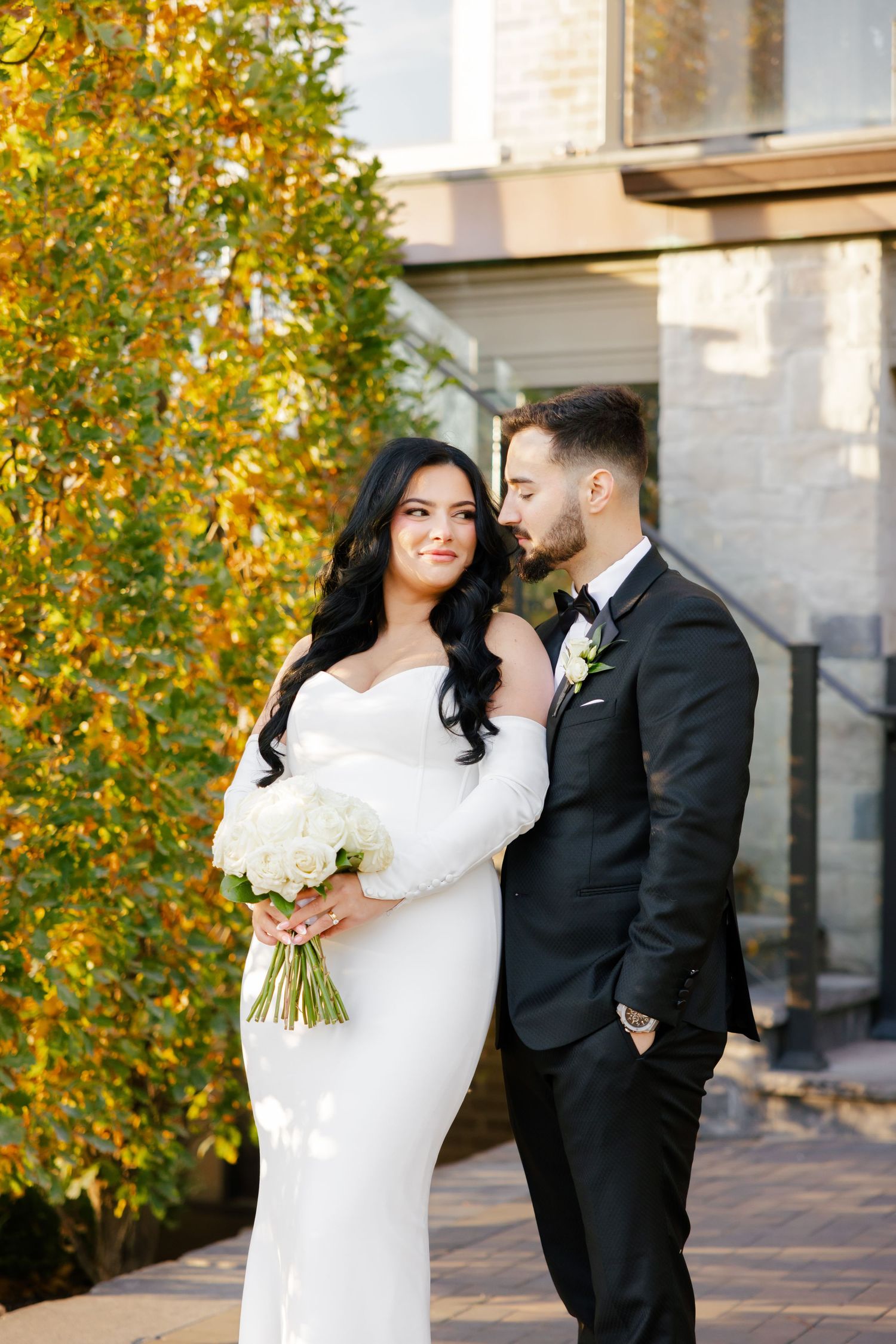 A wedding couple poses against autumn foliage, with the bride in a fitted white dress holding a bouquet of white flowers.