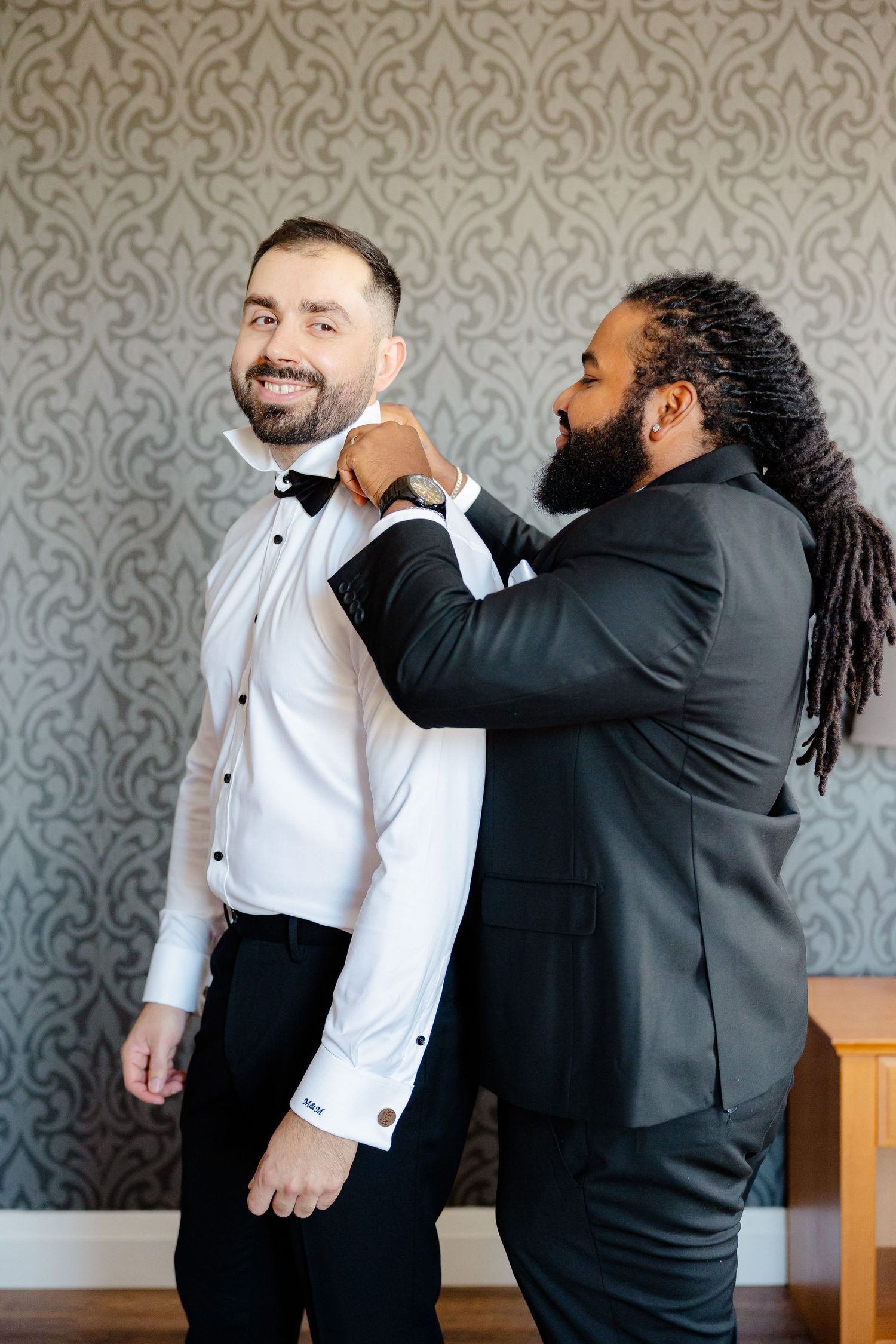 Two groomsmen help each other get ready on wedding day against an elegant damask wallpaper background.