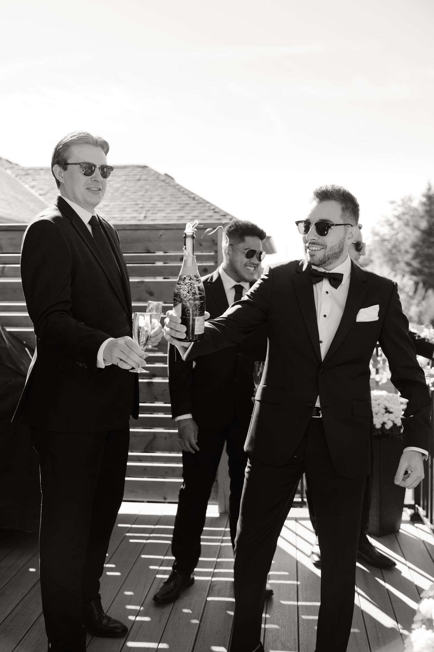 Stylish groomsmen in sunglasses and formal wear socializing with drinks on wooden deck.