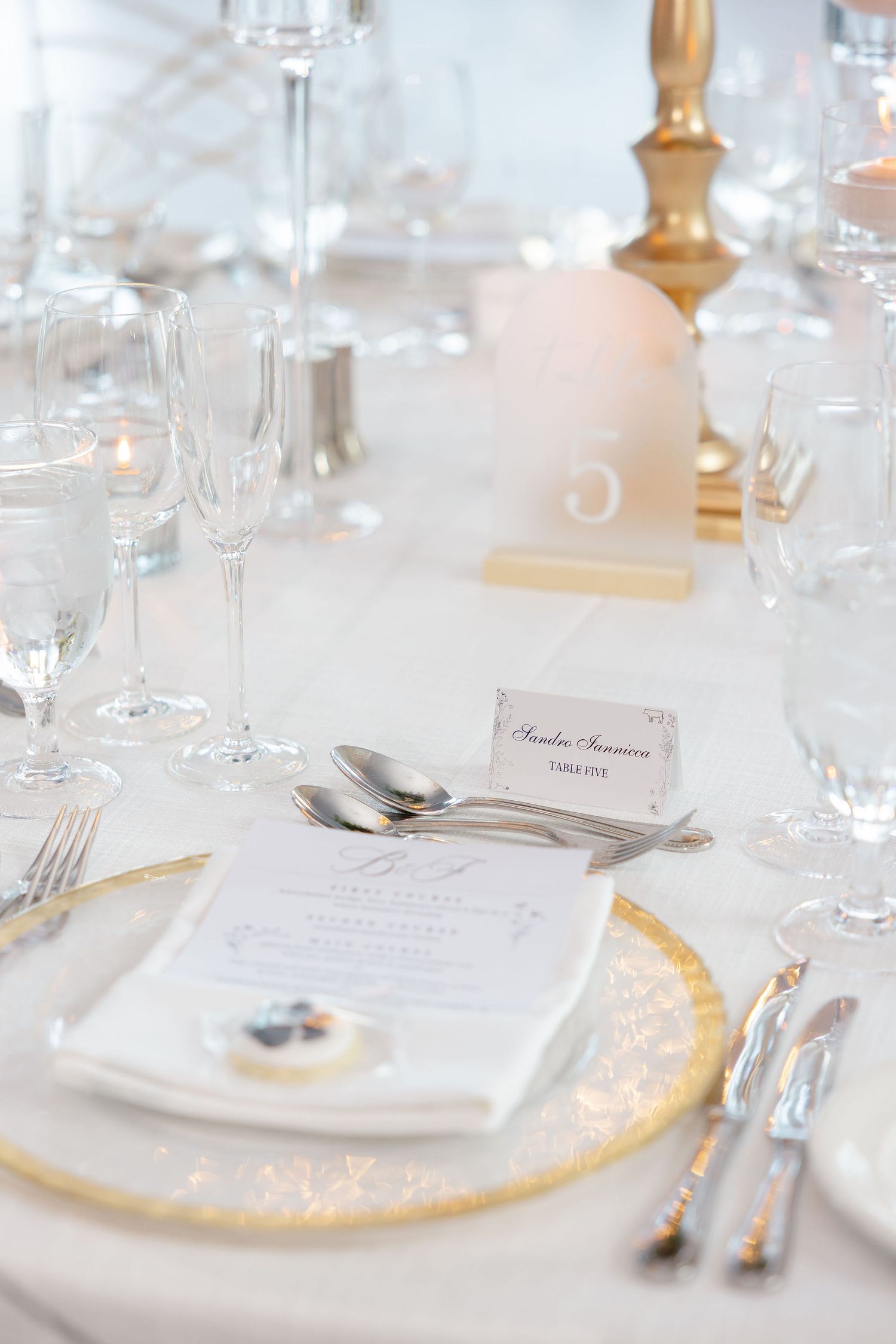 Elegant wedding table setting with white linens, gold charger plates, crystal glasses and menu cards for formal dining.