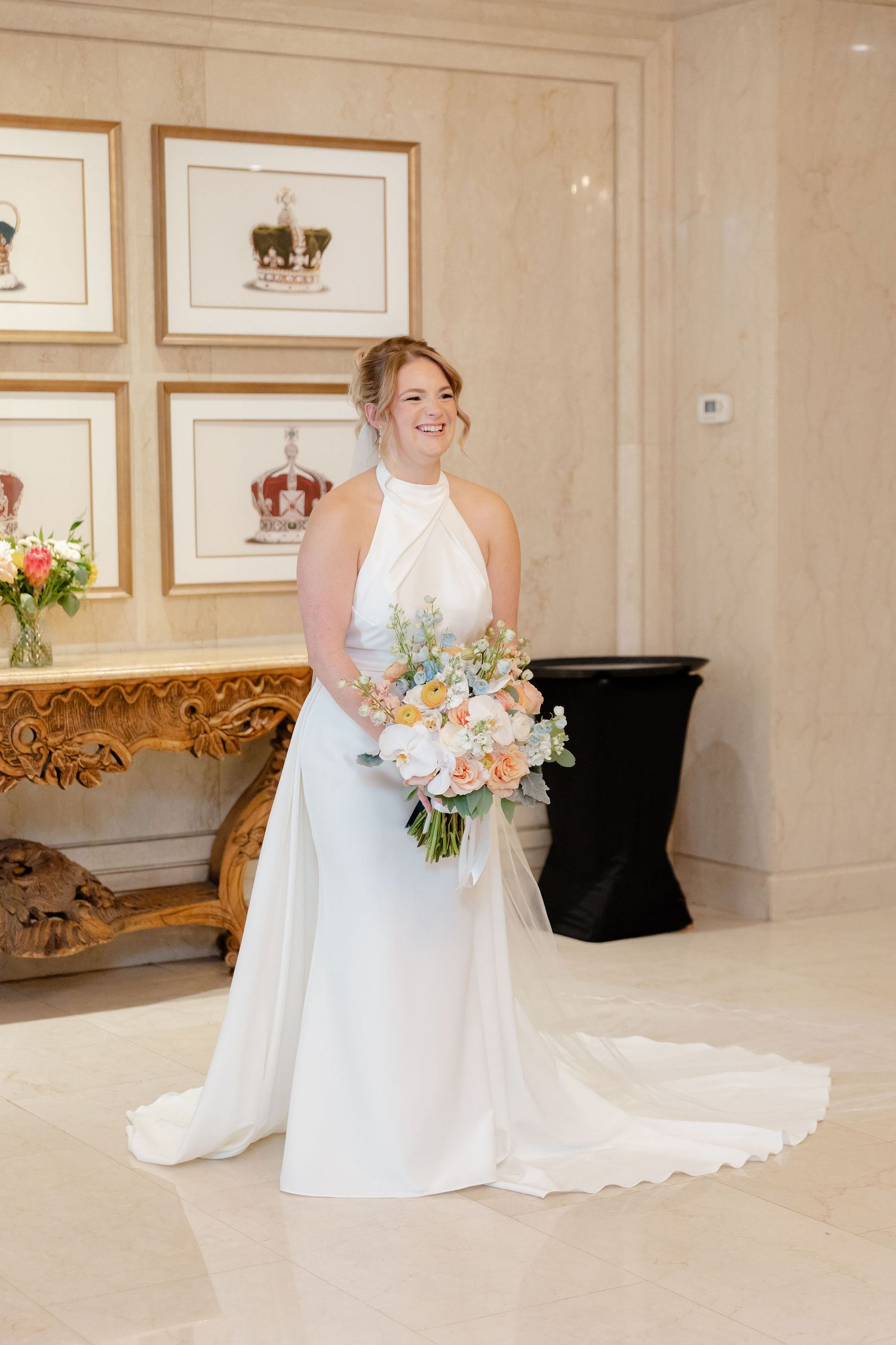 A bride in a flowing white gown poses with a pastel bouquet against an elegant white wall.