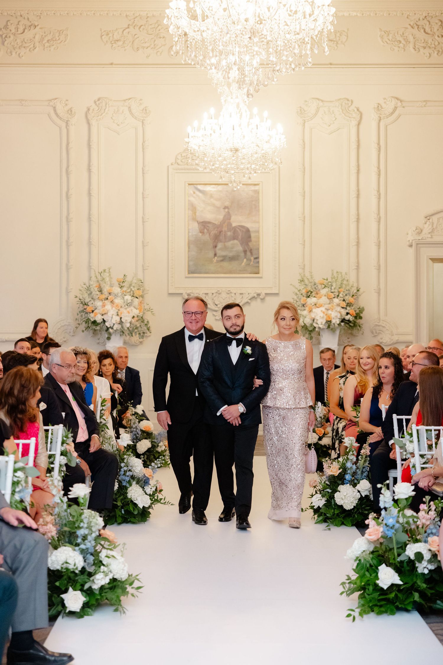 Wedding ceremony processional in an ornate white ballroom with guests seated along the aisle.