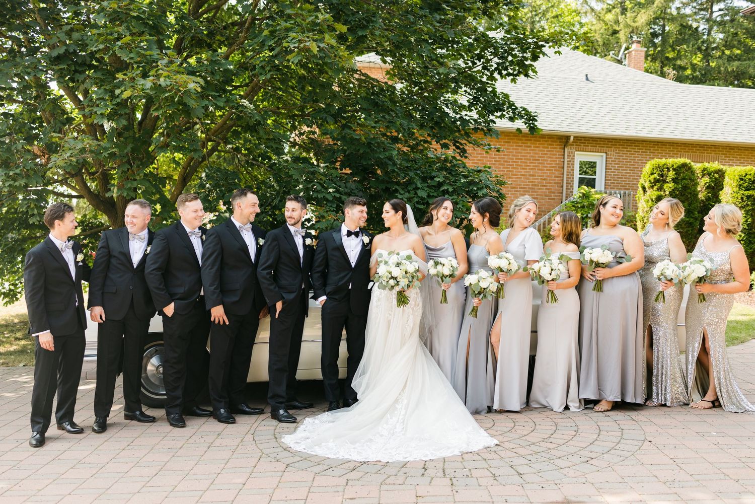 Large wedding party in formal attire arranged for a group photo on a brick courtyard.