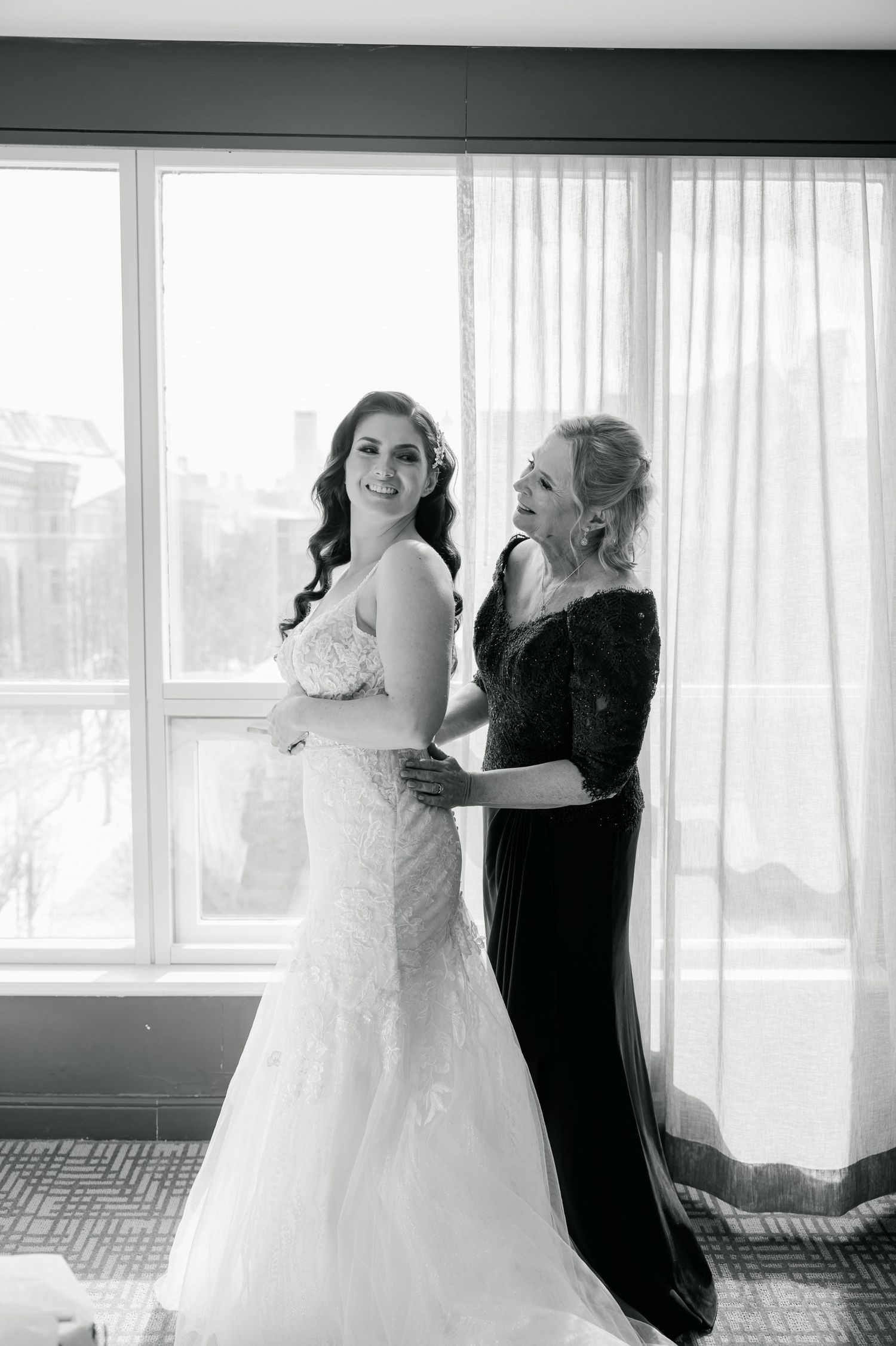 Black and white portrait of bride in lace gown with helper during wedding preparations.