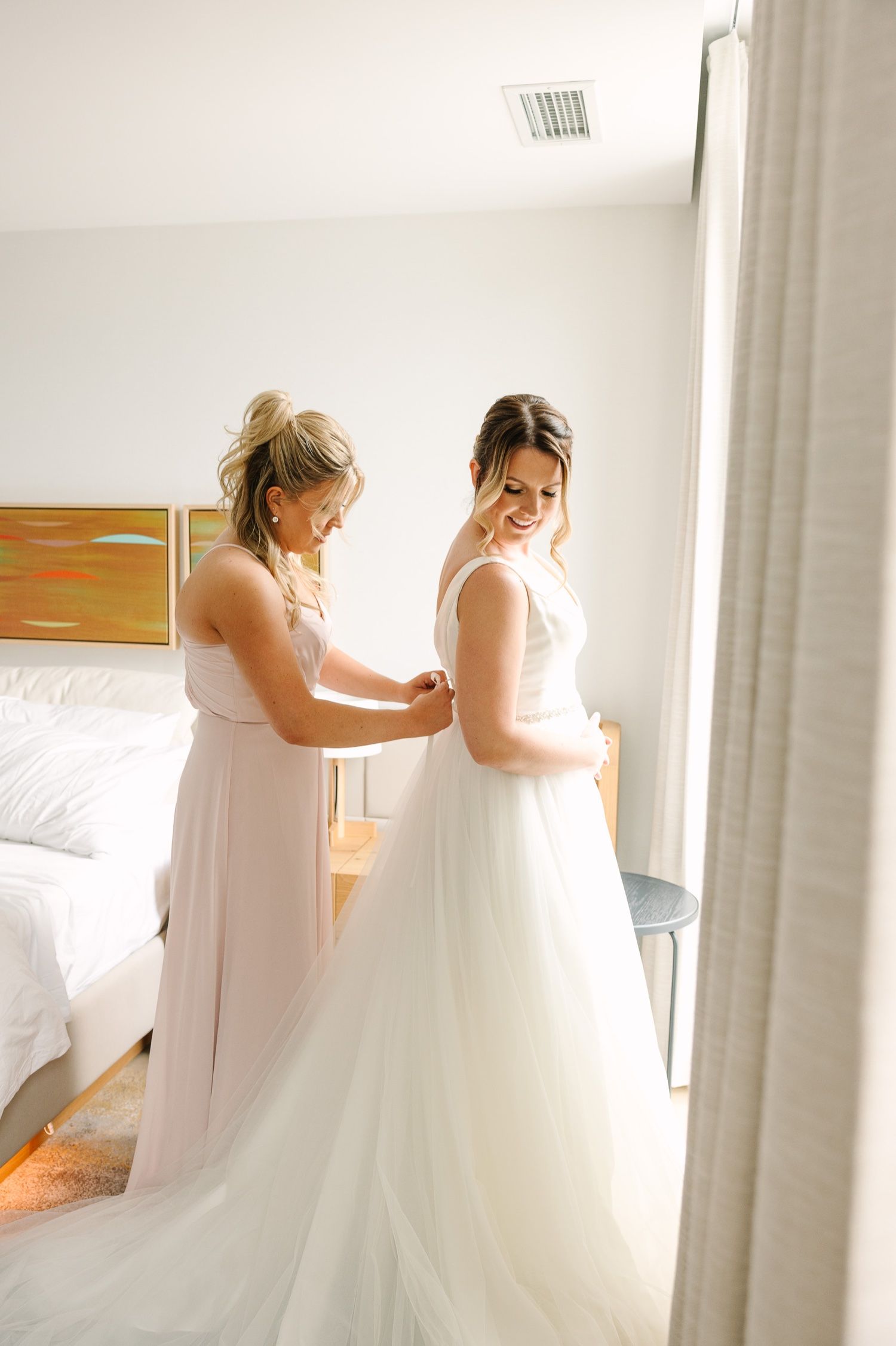 Helper assisting with wedding dress fitting in sunlit room.