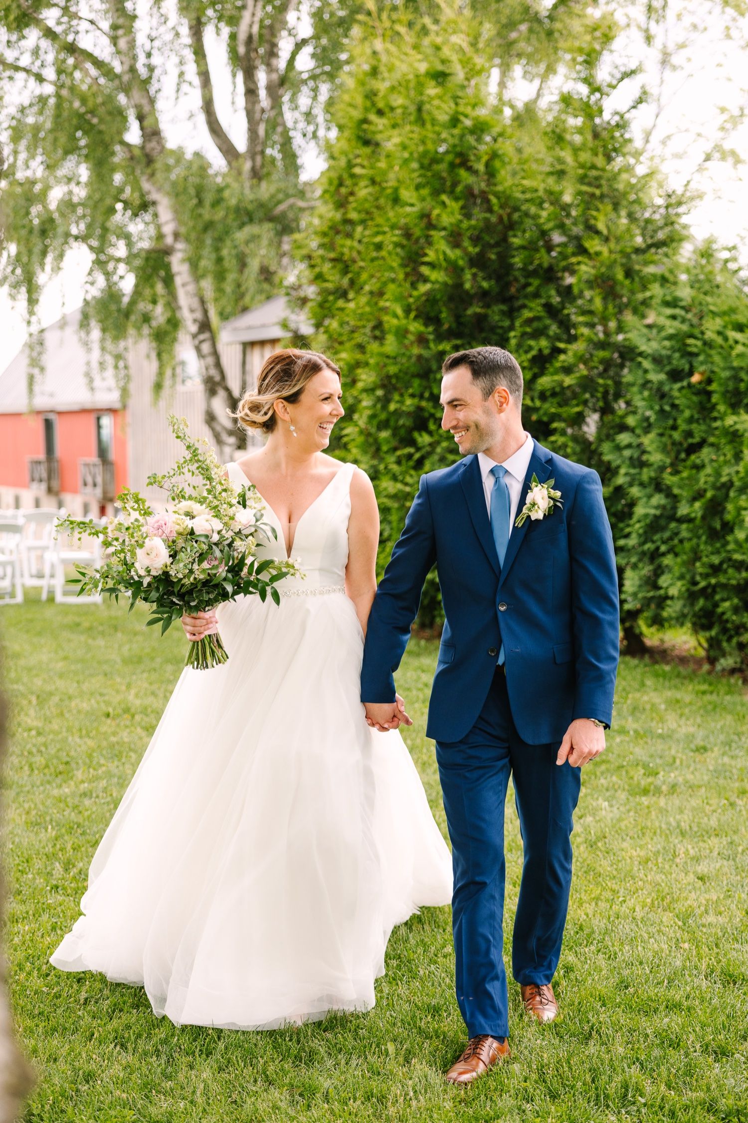 Wedding couple walks hand in hand on grass with elegant white floral bouquet.