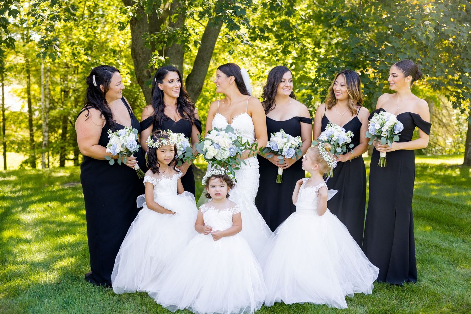 Bridal party with bridesmaids in black and flower girls in white pose in garden setting.