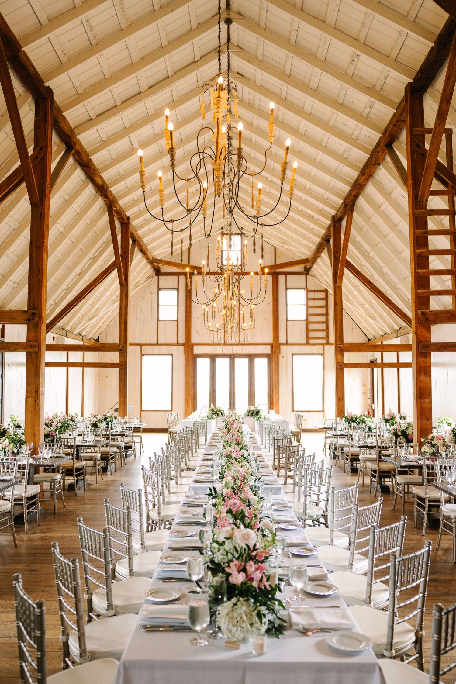 Grand rustic barn interior with vaulted ceiling, chandeliers, and long tables set for a wedding reception.