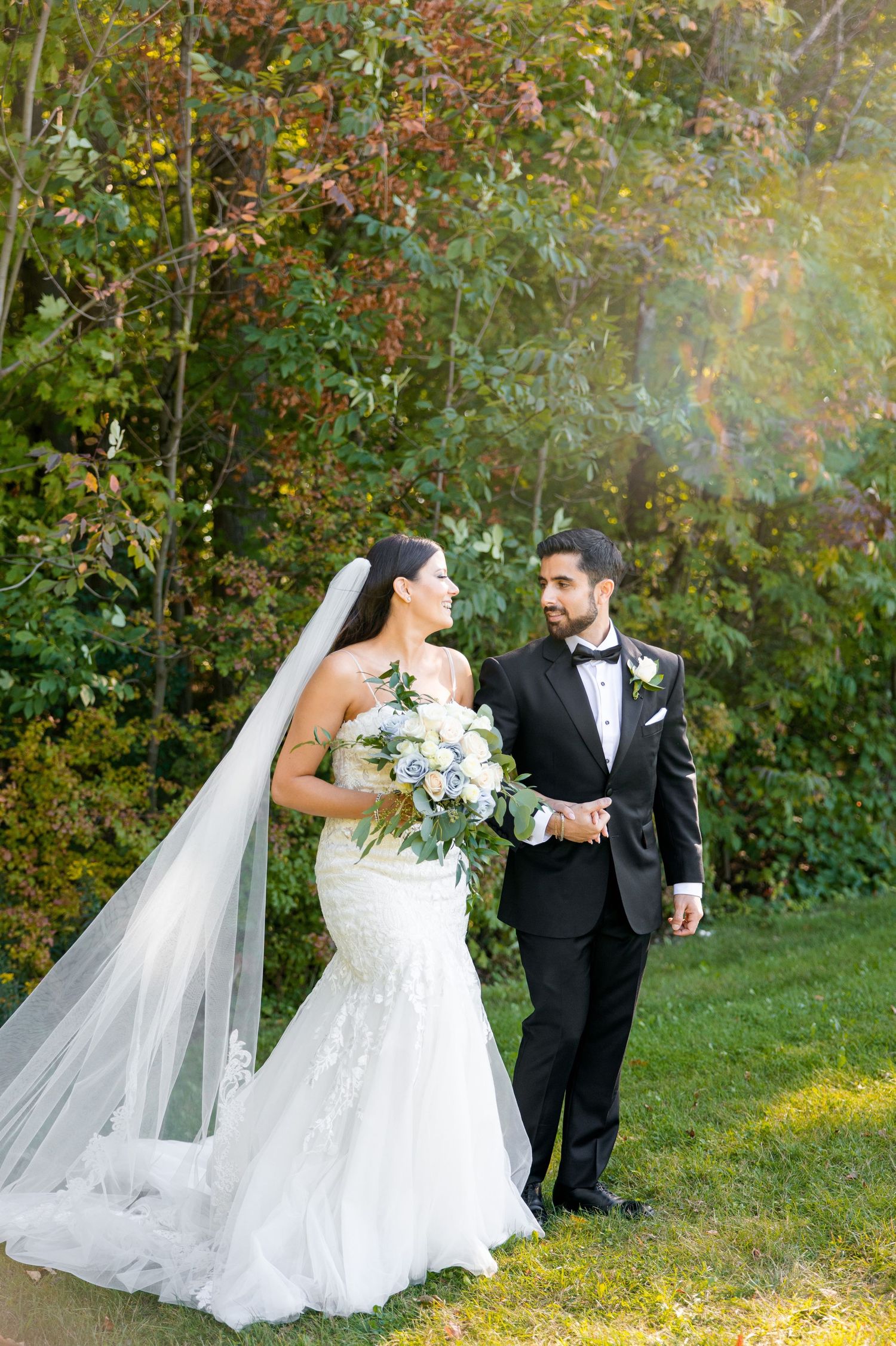 A romantic wedding photo of a couple in formal attire standing outdoors surrounded by autumn foliage.