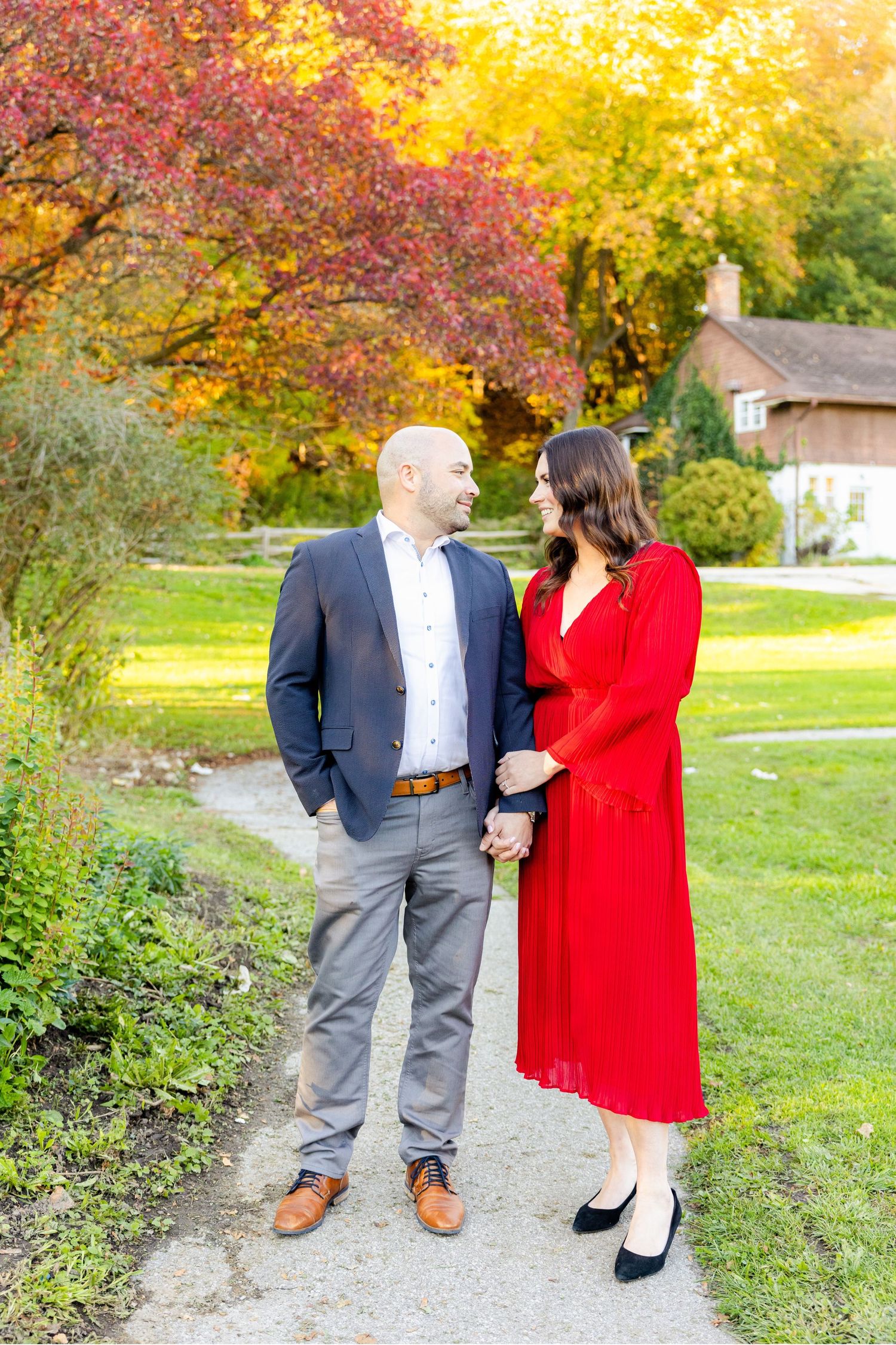 A couple stands together on a garden path with vibrant fall foliage and a house in the background.
