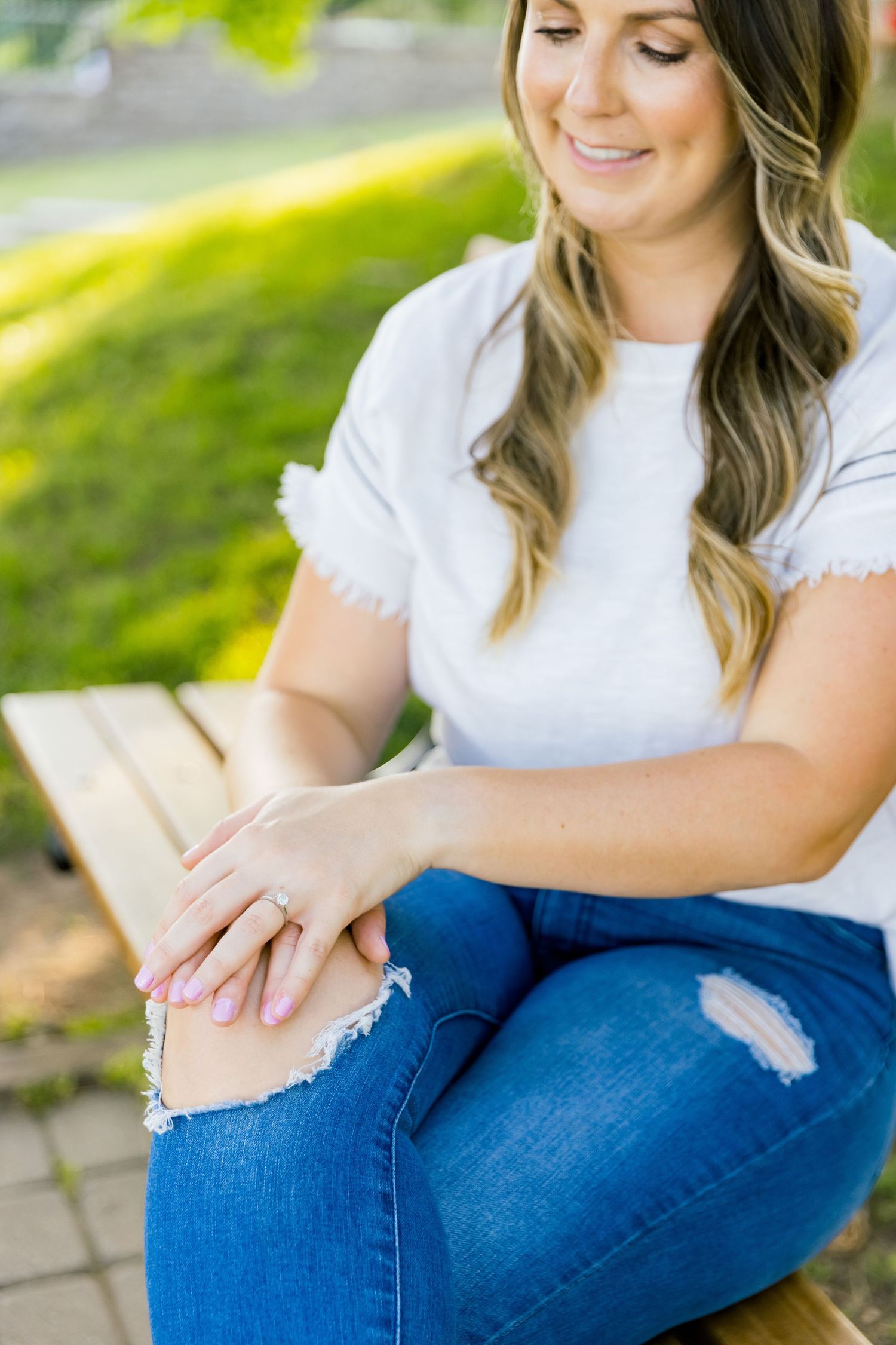 A person in a white top and distressed jeans shows off a ring while sitting outdoors.