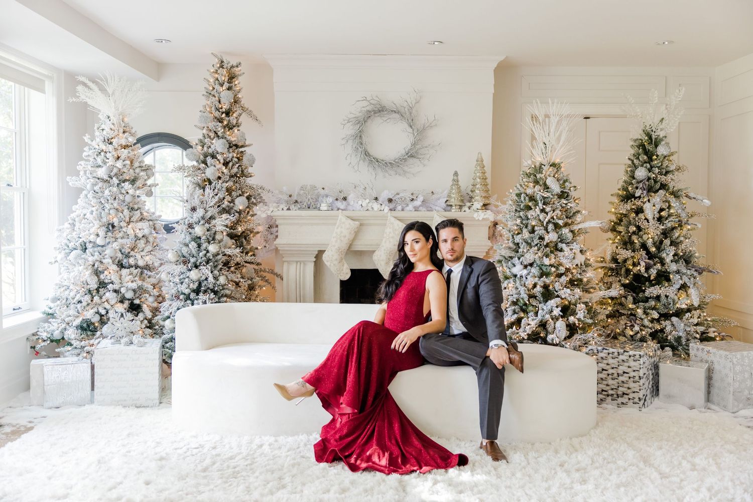 A formal couple sits on white furniture surrounded by decorated Christmas trees and holiday decor.