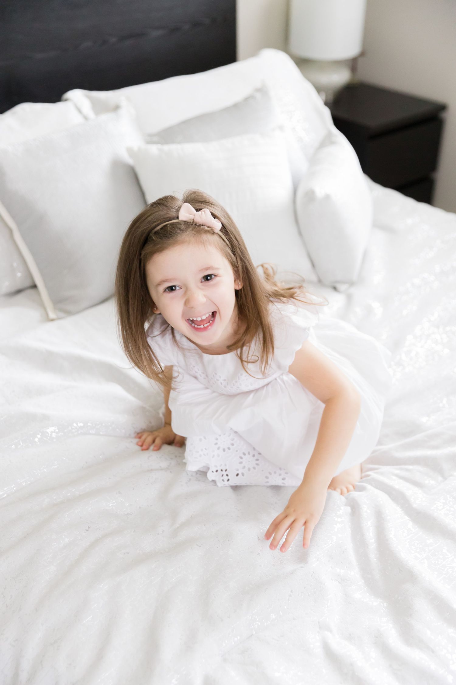 Child in white dress laughing while playing on white bedding.
