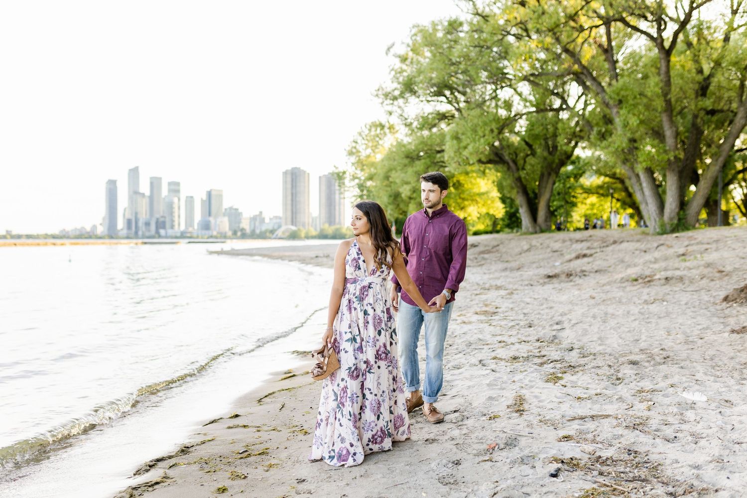 A couple strolls along a sandy beach with a cityscape and trees in the distance.