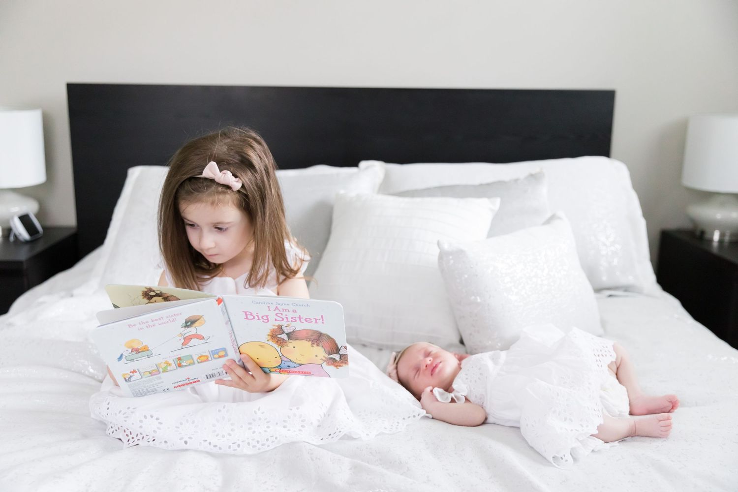 Young girl reading while newborn baby sleeps beside her on white bed.