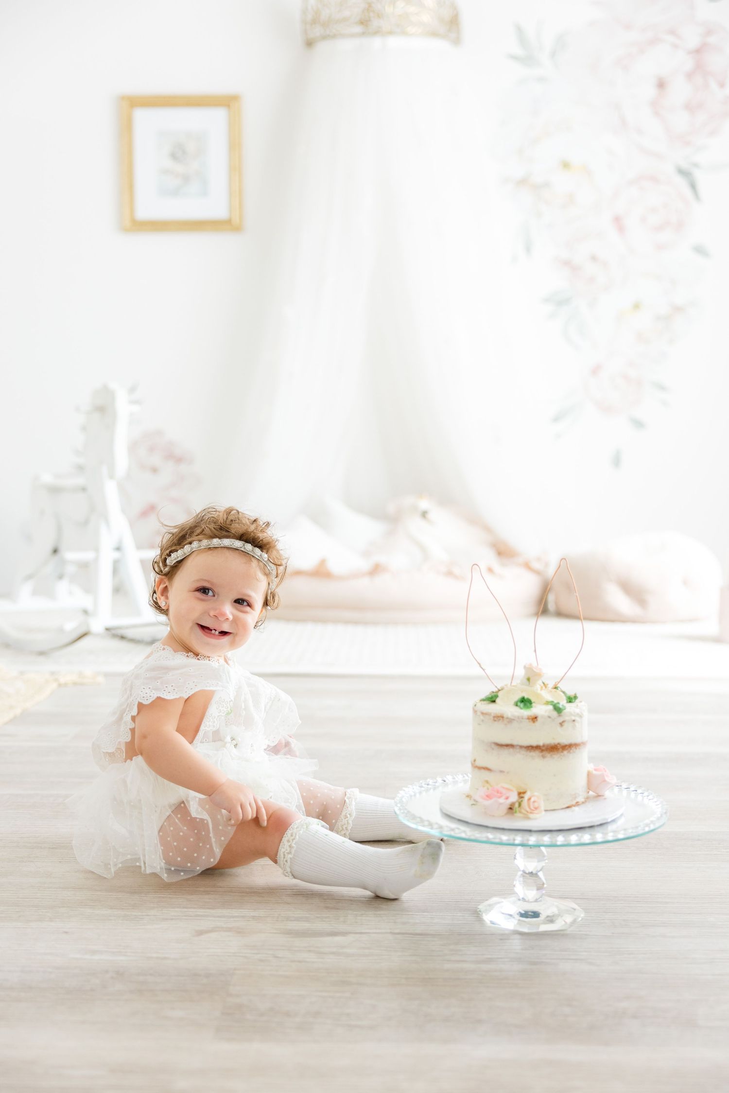 A small child in lace dress smiles while sitting next to a decorated cake with bunny ears on top.