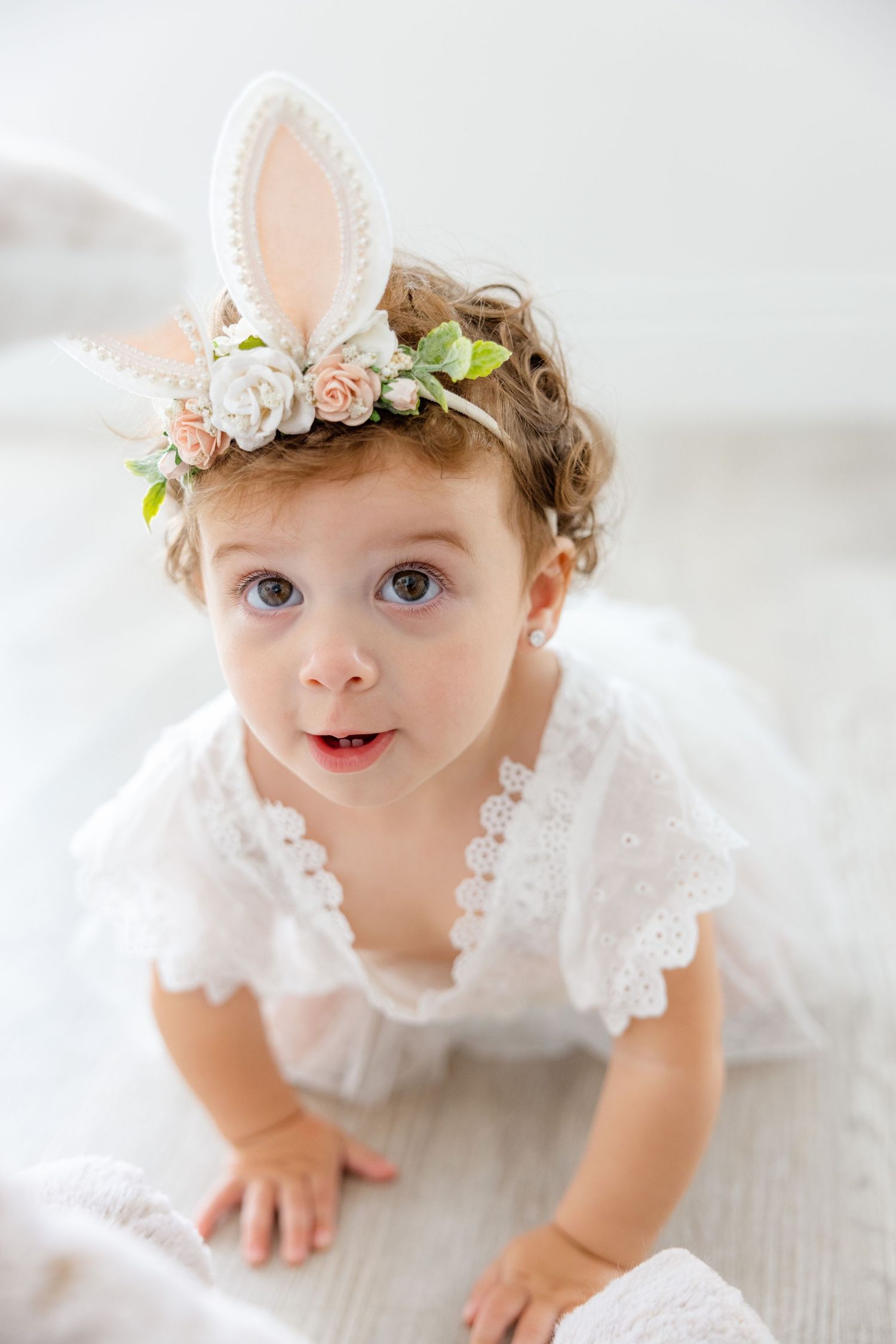 Dreamy portrait in white featuring delicate lace dress and bunny ear crown adorned with flowers.