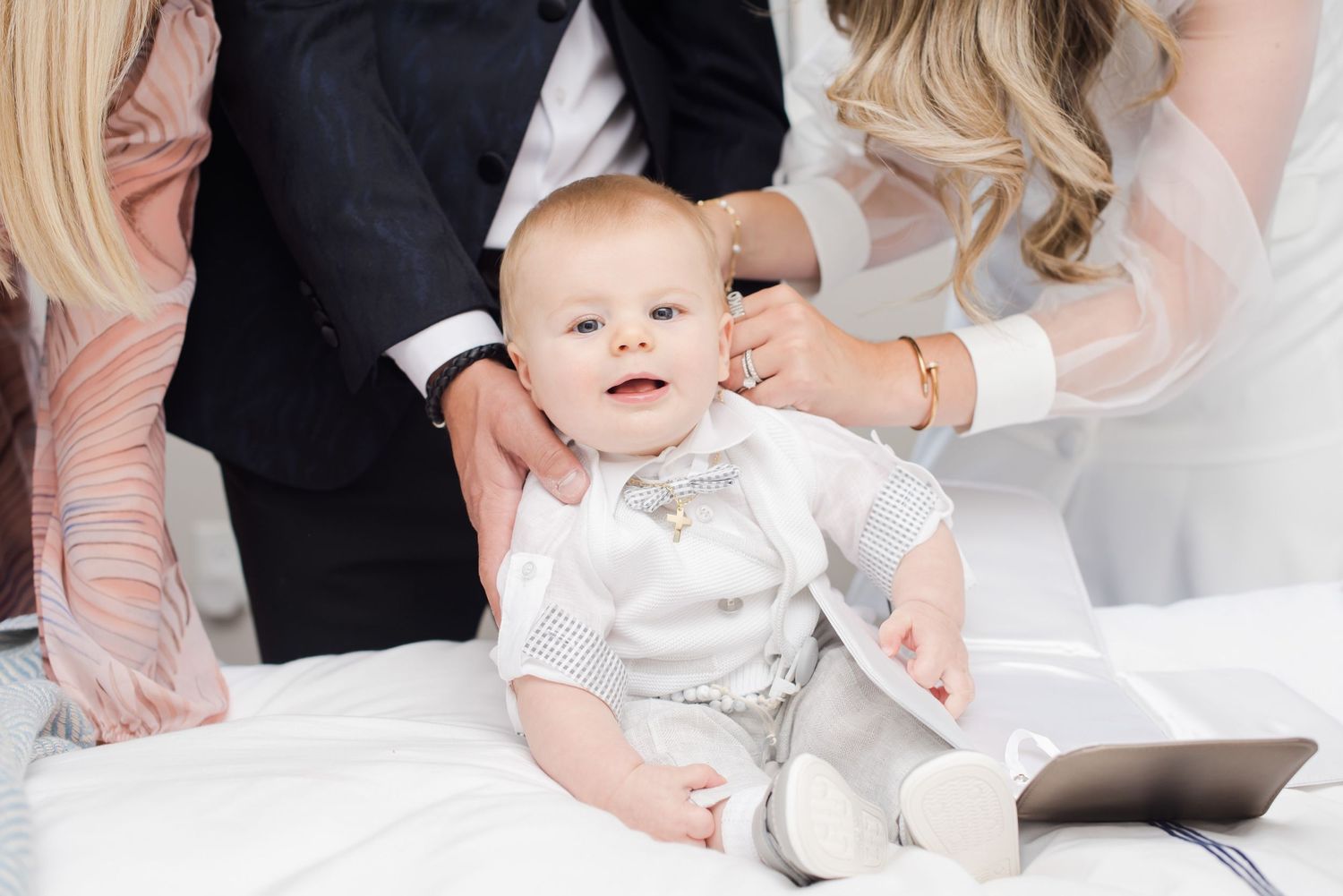 Smiling baby in white outfit sits surrounded by well-dressed adults.