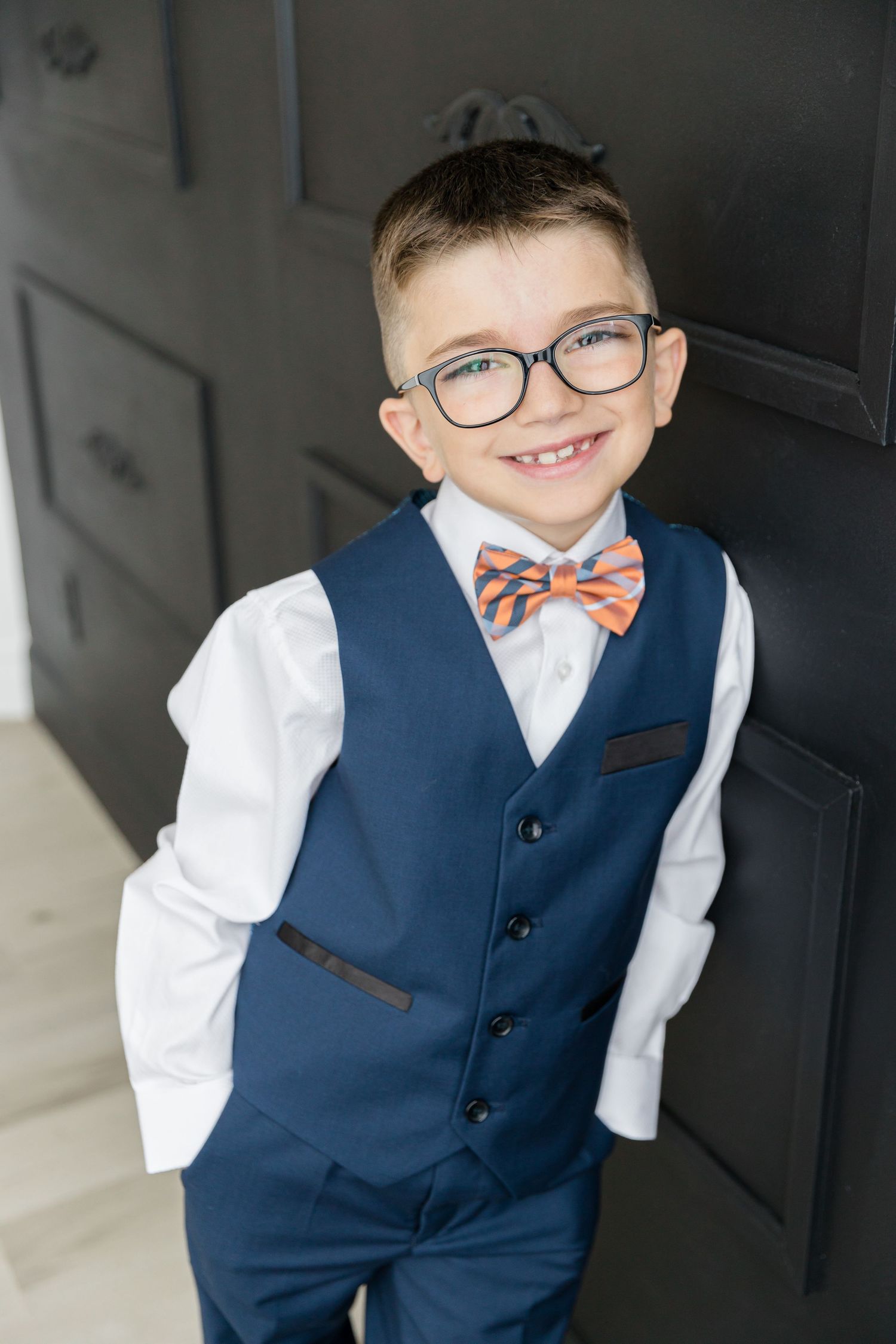 A dapper attire featuring a navy vest, white dress shirt and striped bow tie with glasses against a dark wall backdrop.